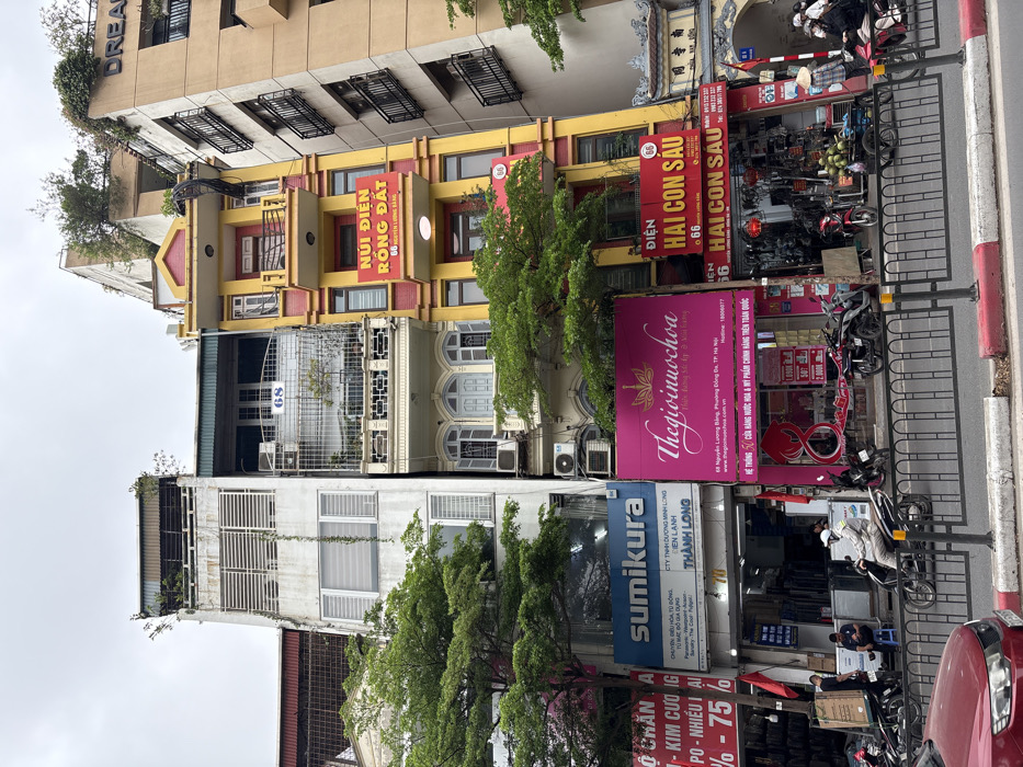 This image depicts a dense urban street scene on Nguyễn Lương Bằng Street, Đống Đa District, Hà Nội, Vietnam, under an overcast sky.
The scene is dominated by a row of multi-story buildings with mixed commercial and residential use. From left to right:
1. A tall, narrow white building (partially obscured) with a vertical vine, horizontal louvered windows on its upper levels, and a balcony with an iron railing and vegetation on its uppermost floor, marked with the number "68". Its ground floor houses a "sumikura" electronics store and a business named "THÀNH LONG".
2. Adjacent to it, another narrow building, also implicitly at "68", features a light-colored, more ornate facade with arched window frames and a balcony with plants on an upper floor. Its ground floor is occupied by "thegioinuochoa", a perfume store with a prominent pink sign.
3. Next is a distinct multi-story building painted vibrantly in yellow with red trim and featuring multiple red-railed balconies. Signs on its facade indicate "NÚI ĐIỆN RỒNG ĐẤT" on an upper level and "ĐIỆN HAI CON SÁU" (an electronics store) on the ground and first floors, both displaying the address "66 Nguyễn Lương Bằng".
4. To the far right, a taller, light brown building with multiple dark-railed balconies is partially visible, with the word "DREAM" discernible near its top.
At street level, a black metal barrier with red and white striped bases separates the street from the sidewalk area. Numerous motorbikes are parked along the sidewalk. A person wearing a white jacket and helmet is actively riding a motorbike from left to right in the mid-ground. Near the "sumikura" store, two individuals are visible, one seated. Closer to the "ĐIỆN HAI CON SÁU" store, a street vendor wearing a conical hat (nón lá) stands beside a small cart laden with coconuts. Other pedestrians and parked motorbikes are present in the vicinity of the shops. The front fender of a red car is visible in the immediate foreground, positioned on the roadway. Green trees with dense foliage are interspersed along the street, partially obscuring the lower and mid-sections of the buildings.