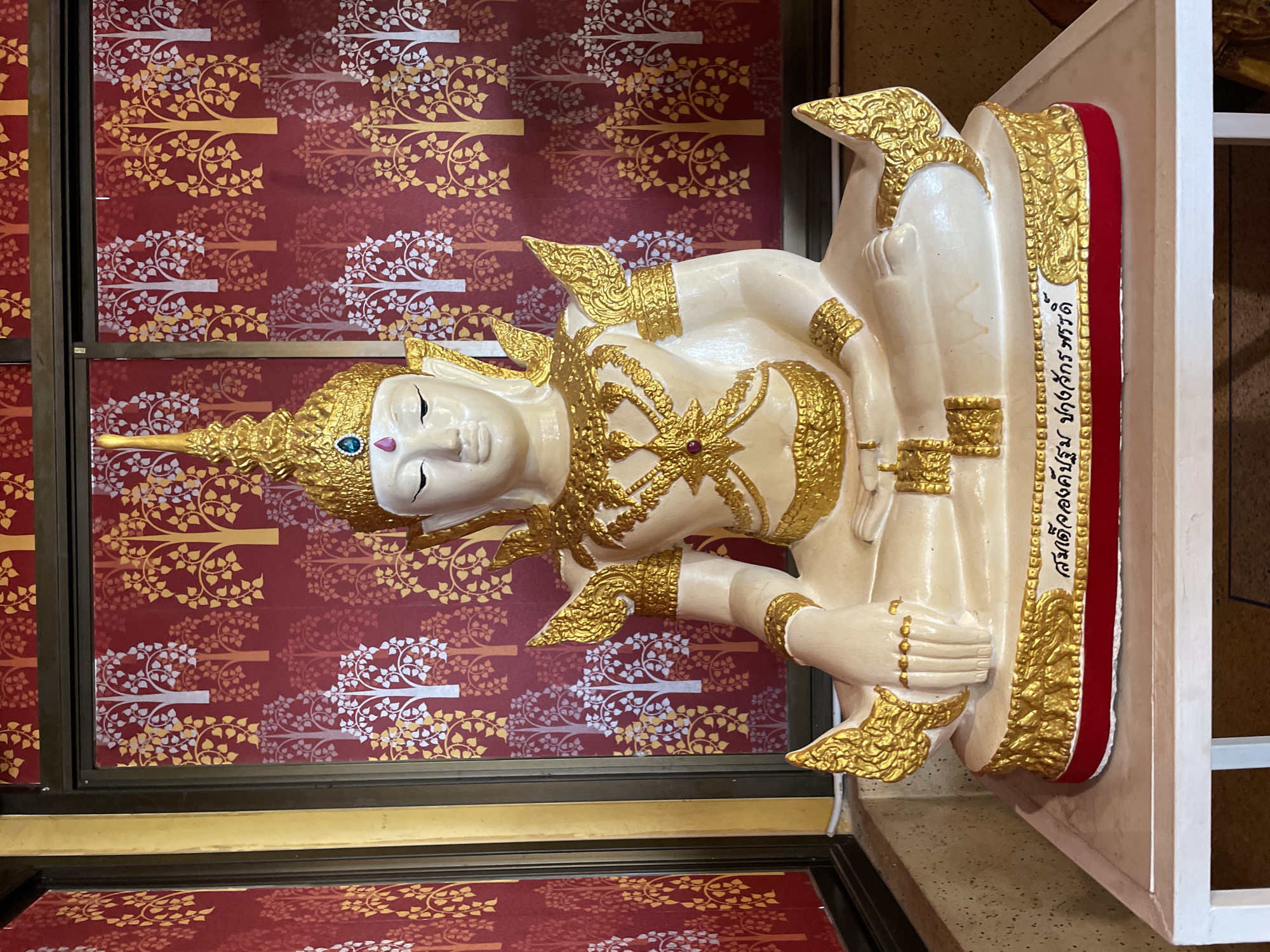 This is a frontal, eye-level, medium shot of a white and gold Buddha statue seated in meditation. The statue is ornately decorated with gold trim, jewels on its headdress and chest, and gold leaf accents on its shoulders and knees. The statue is positioned in front of a red wall with a repeating pattern of stylized trees in white and gold. The base of the statue is also gold and features Thai script. The statue is placed on a white platform with a red trim, which is itself on a larger white surface, possibly a table or altar. The background appears to be an interior space, possibly a temple or shrine in Bang Sare, Thailand, given the style of the statue and the script.