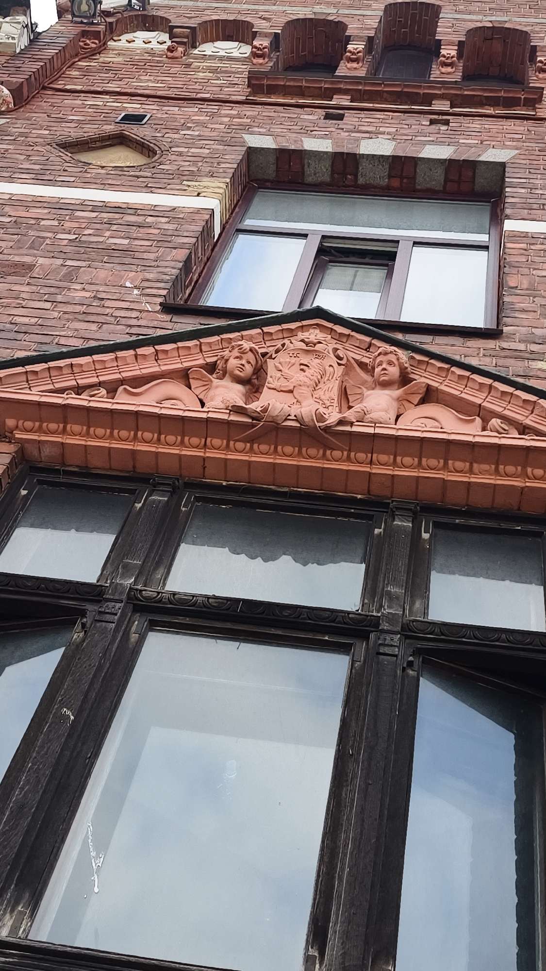 The image shows an upward angle of a brick building facade in Vyborg, Russia. The building features red brickwork with decorative elements, including a pediment above a window with two sculpted cherubs. The window below the pediment is divided into multiple panes with dark wooden frames. The overall architectural style suggests historical significance, potentially from the late 19th or early 20th century. The sky is visible through the window panes, reflecting a light gray, overcast day. No people are visible in the image.