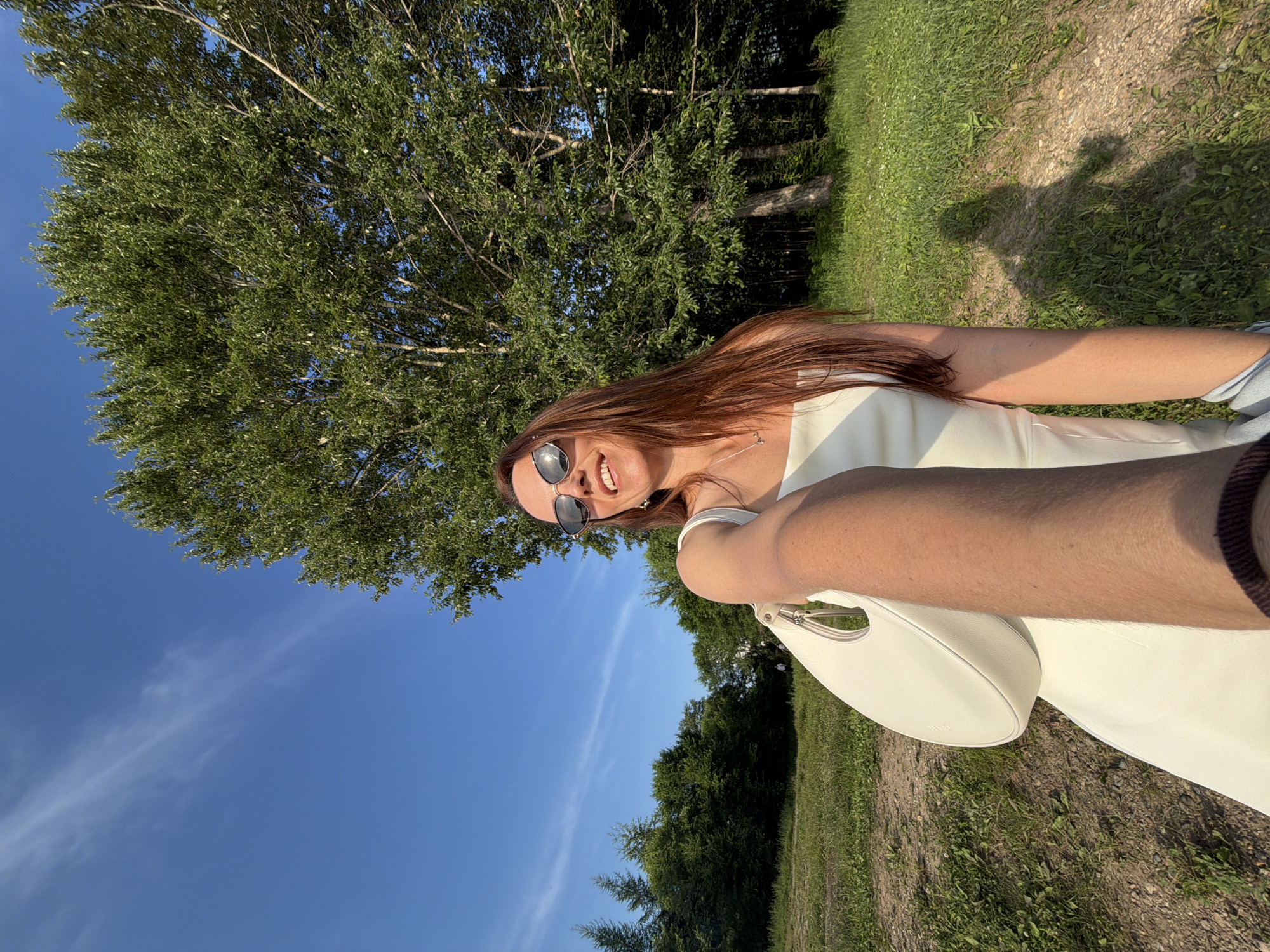 A woman is taking a selfie outdoors, with a large tree and a blue sky in the background. She is wearing a white dress and sunglasses, and holding a white handbag. The woman has long brown hair and is smiling. The sun is shining, casting shadows on the grass. The location is the city of Birobidzhan, Russia.