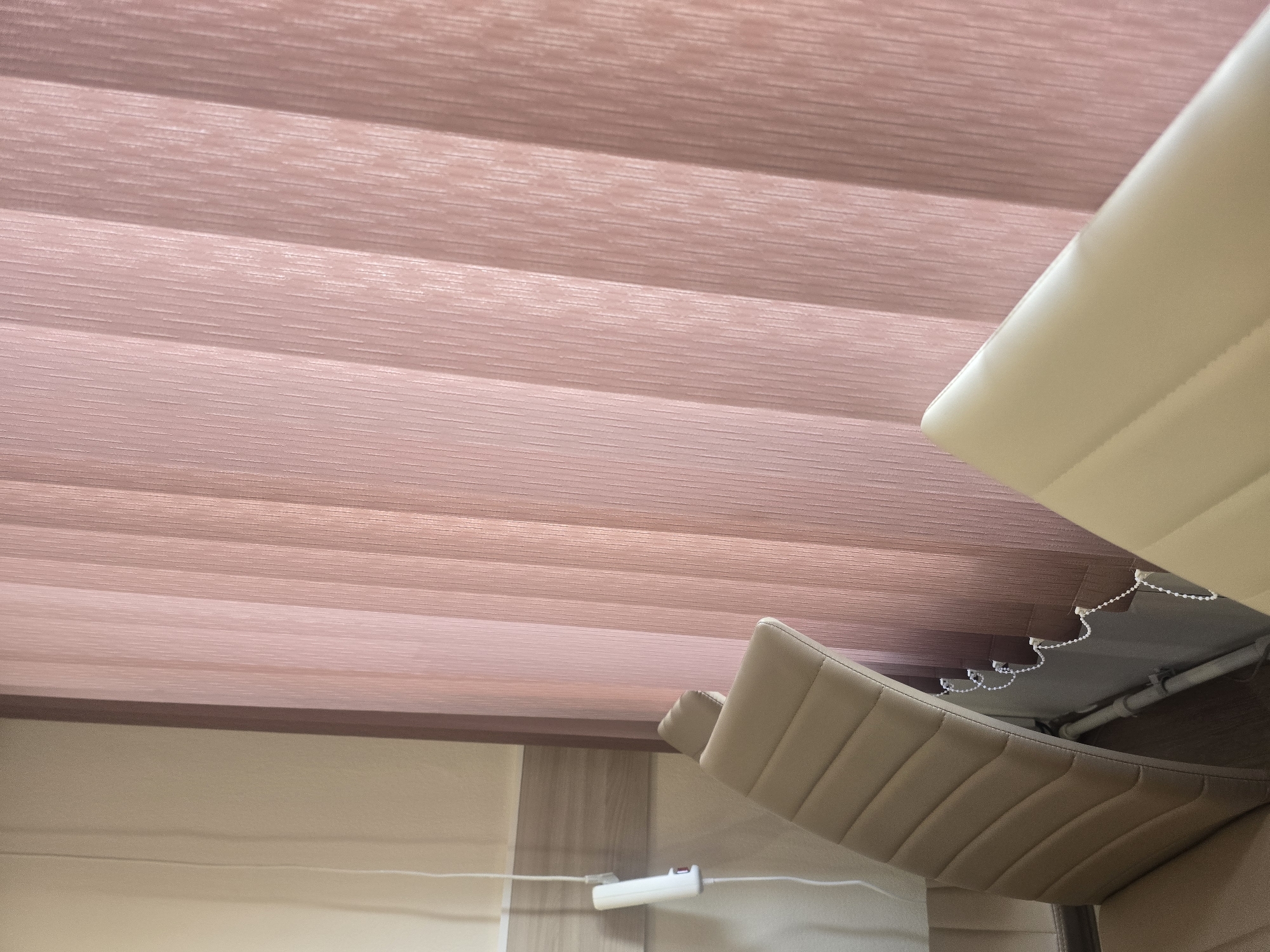 The image shows a close-up view of vertical blinds in a pale pink color, with vertical lines creating a textured pattern. To the left of the blinds, the top of a beige upholstered chair is visible, showing a quilted design on the backrest. Further to the left and out of focus, a white object with a cord is mounted on a light-colored wall with a horizontal wooden trim. The overall scene appears to be indoors, possibly an office or waiting area. The image does not contain any elements that would allow for a determination of its location within Ulan-Ude, Russia.