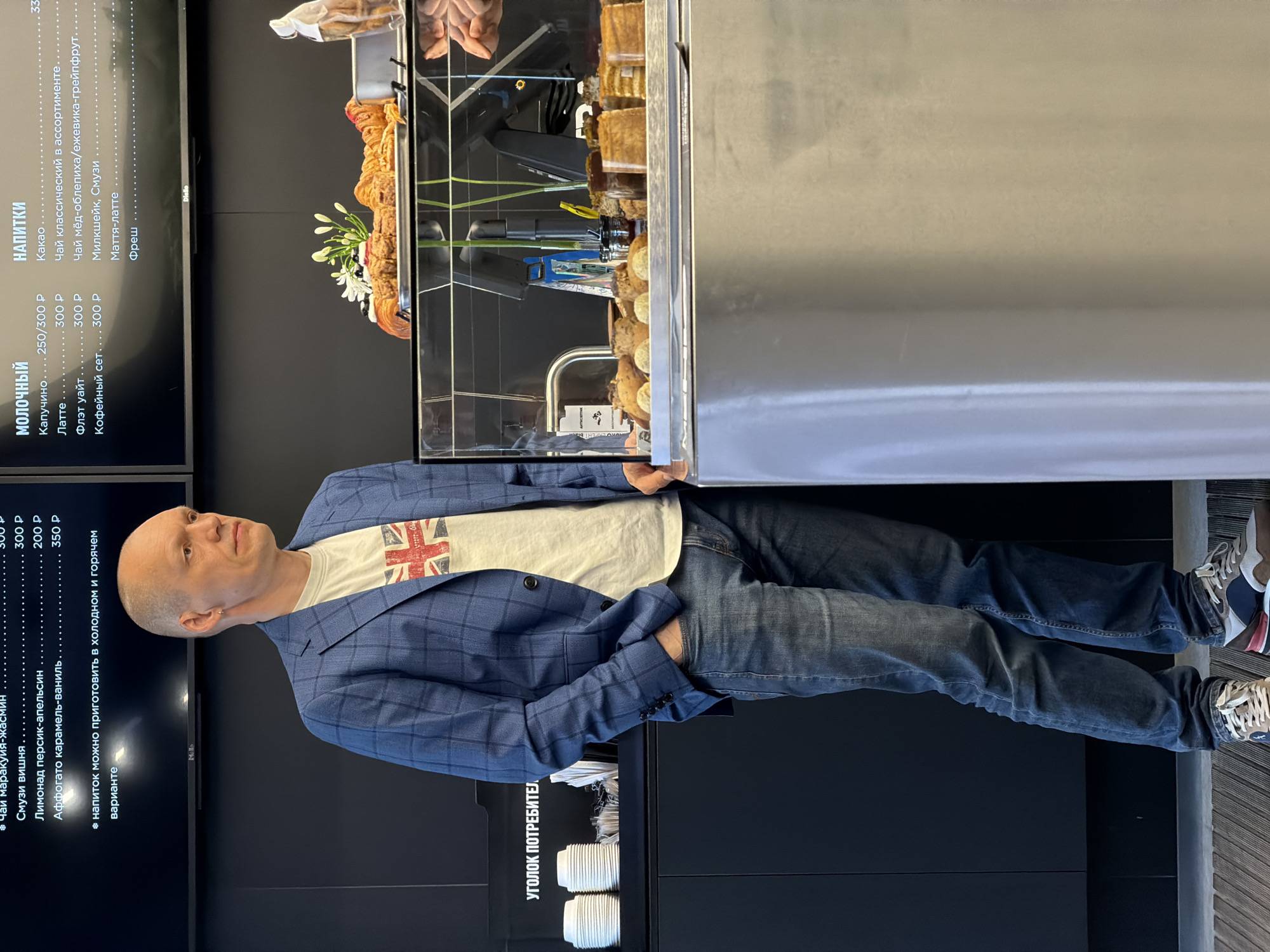 A man stands at a pastry display case in Izhevsk, Russia. He is wearing a white t-shirt with a Union Jack on it, a blue plaid blazer, and blue jeans. He has his hand in his pocket. Behind him are the menu boards for the cafe in Russian. There is a stack of disposable cups in a corner of the counter near the bottom of the shot. Pastries are visible in the display case.