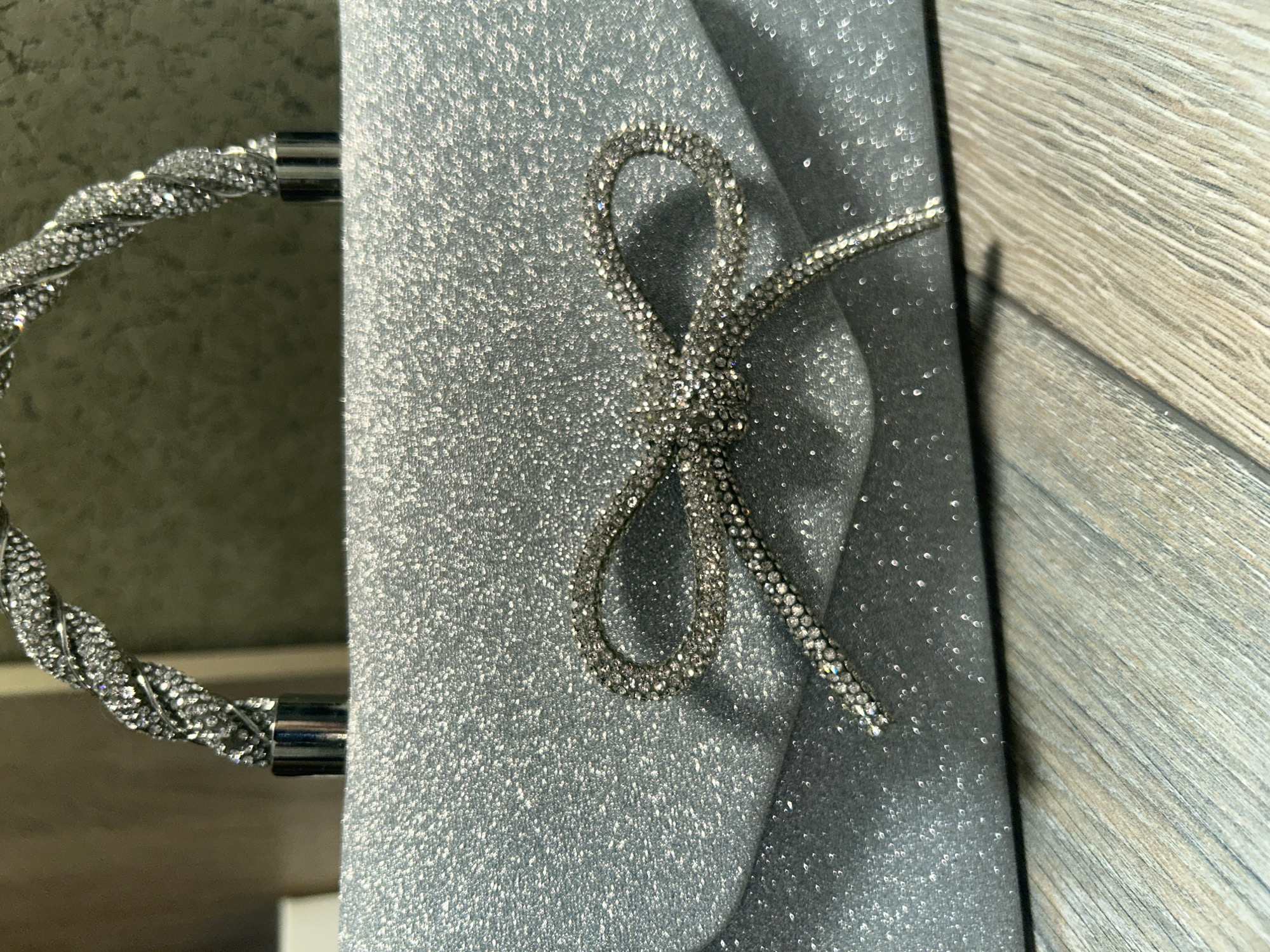 The image shows a close-up of a sparkly silver clutch bag with a bow made of rhinestones. A twisted silver chain strap with rhinestone accents is attached to the bag. The bag is placed on a surface that appears to be made of wood planks with a greyish-brown tone. The lighting suggests it is daytime, possibly indoors, as there are subtle shadows and reflections from the rhinestones and shiny surfaces. The overall aesthetic is glamorous and festive, suggesting it could be an accessory for a special occasion. There is no visible text in the image. The setting is likely a domestic or retail environment where such an accessory would be displayed or used.