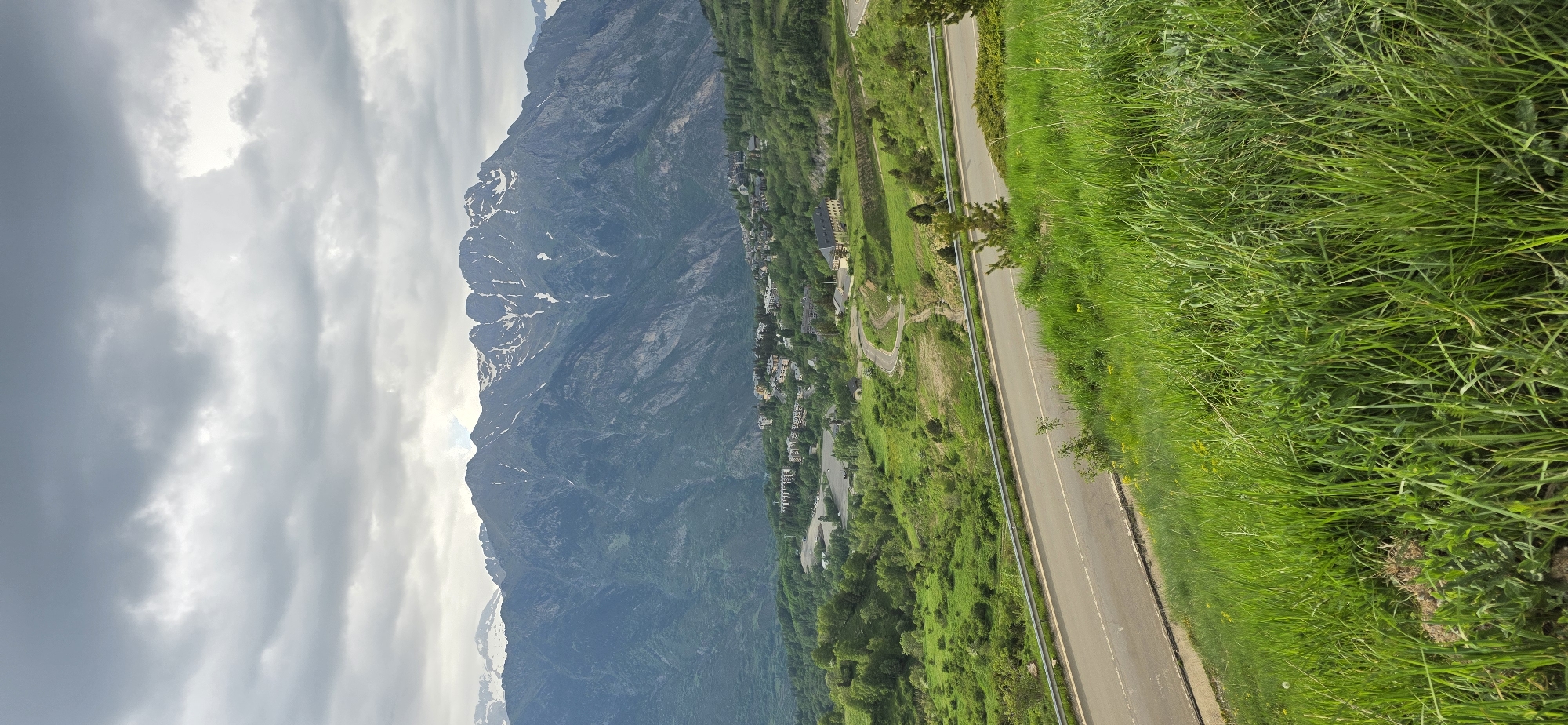 The image depicts a mountainous landscape in Benasque, Spain. A steep mountain range with patches of snow on its upper slopes dominates the left side of the frame, partially obscured by cloudy skies. A road runs parallel to the mountain, lined with lush green grass and vegetation. A town or village is nestled between the road and the base of the mountain, consisting of numerous buildings.