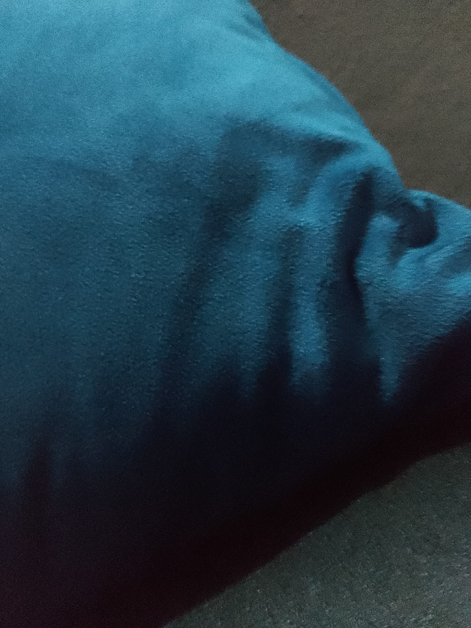 The image shows a close-up view of a crumpled teal-colored pillow, likely made of a soft fabric with a slight textured surface. The lighting is dim, casting shadows and highlighting the folds and creases of the pillow. The background is dark and out of focus, suggesting a dimly lit room. No specific location in Jauro Yinu, Nigeria can be determined from this image.