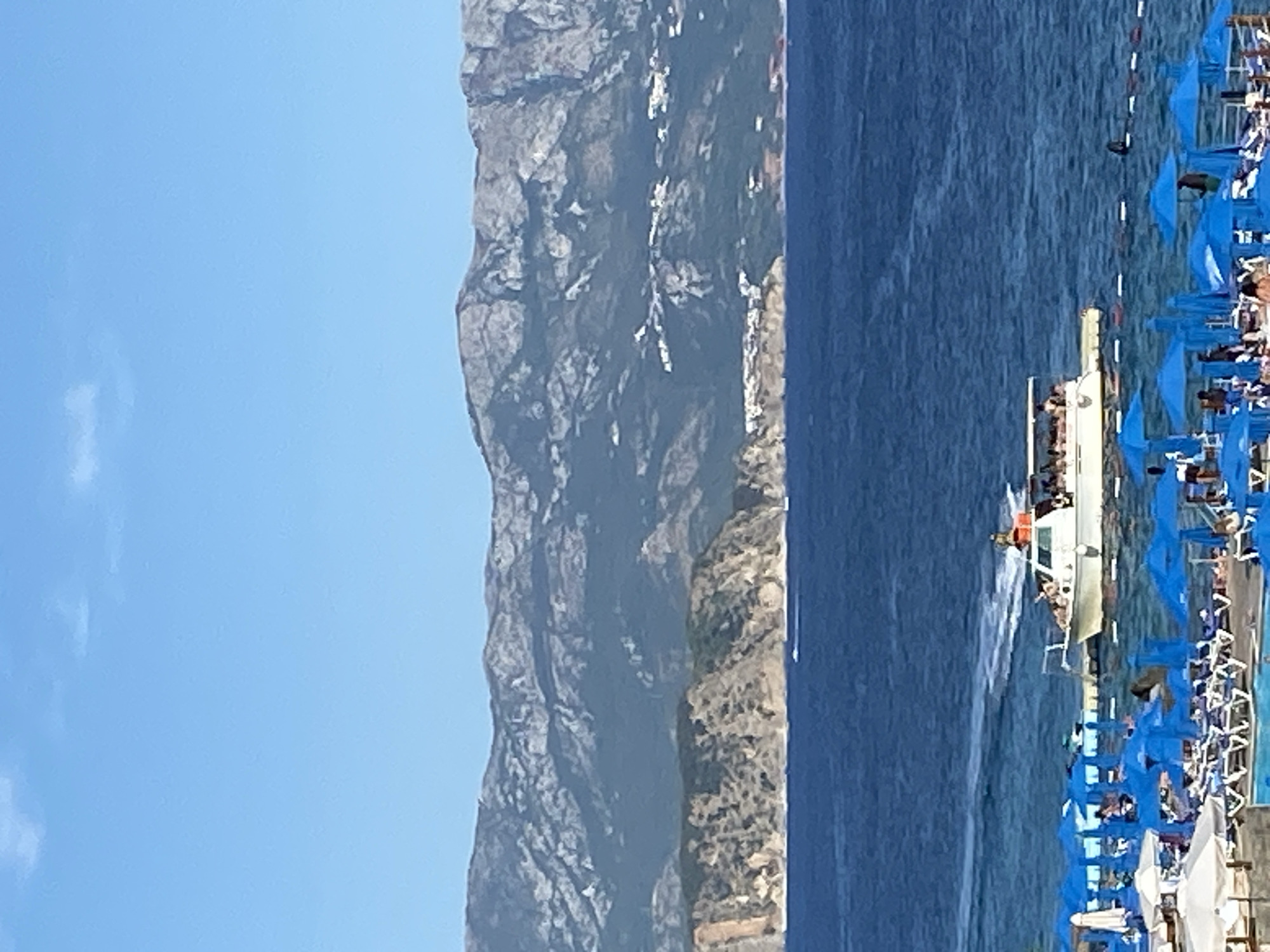 A bright, sunny day scene depicts a coastal view with mountains in the background and a beach in the foreground. The azure sea stretches to the horizon, meeting a clear blue sky. In the middle distance, a white boat with a canopy is navigating the water, carrying passengers. The boat leaves a white wake behind it. The coastline features rugged, rocky cliffs dotted with sparse vegetation, and some structures are visible on the slopes of the distant mountains.
In the foreground, a beach is set up with numerous blue umbrellas and lounge chairs arranged on the sand. Some people are visible under the umbrellas and on the beach, with others possibly in the water. A white structure, possibly a building or a large umbrella, is also present on the beach. The scene suggests a popular tourist destination, likely a resort area. The image captures a moment of leisure and recreation on a warm day by the sea, with the dramatic mountain landscape providing a striking backdrop.