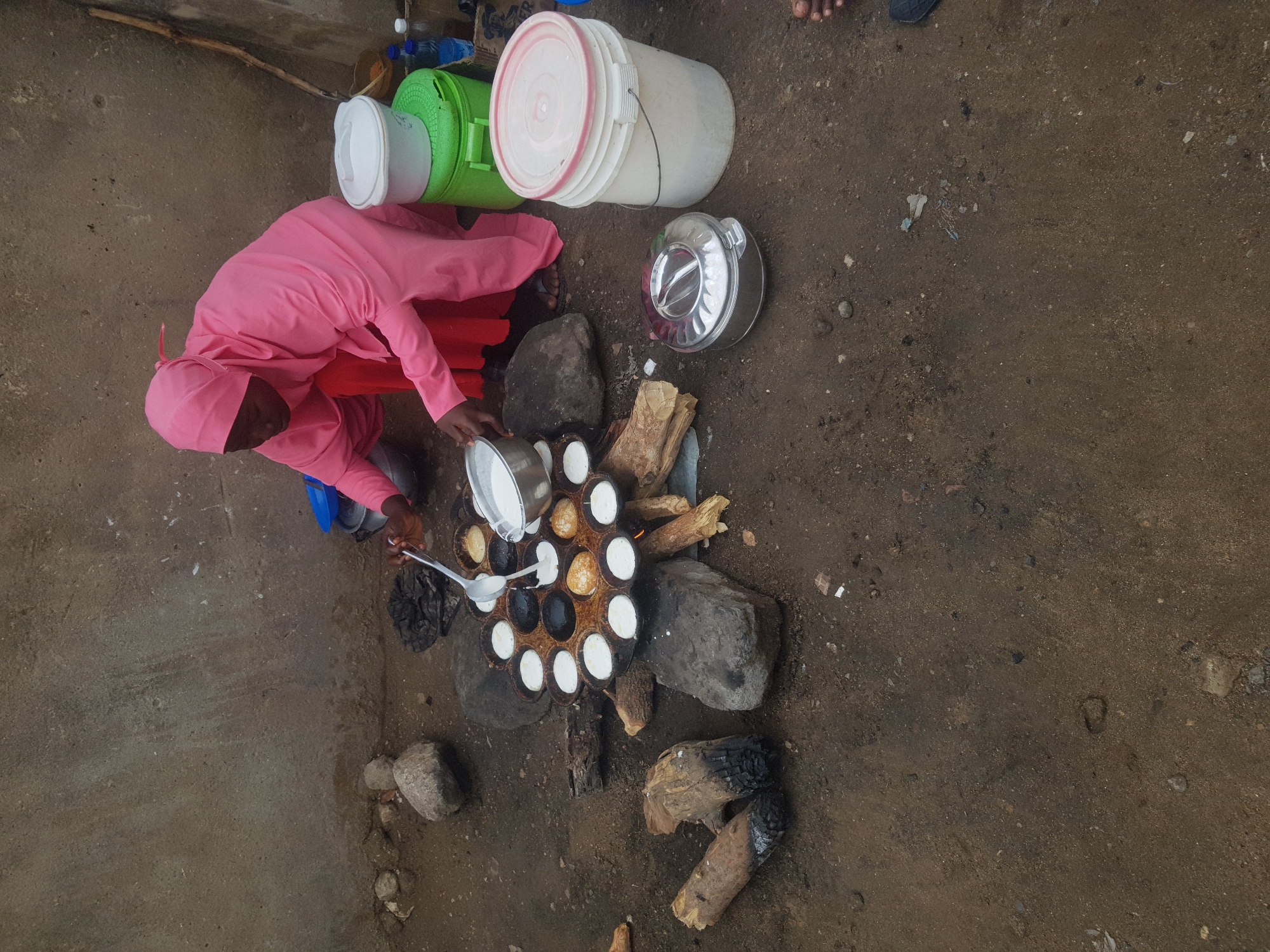 A person in a pink robe and head covering is cooking on the ground in Bauchi, Nigeria. The cooking setup consists of a round metal pan with multiple circular indentations, surrounded by stones and fueled by wood. The person is pouring a white liquid from a metal container into the indentations of the pan using a spoon. There are several buckets, a shiny insulated food container, and various cooking utensils nearby. The ground is bare earth. A concrete wall is visible on one side of the scene.