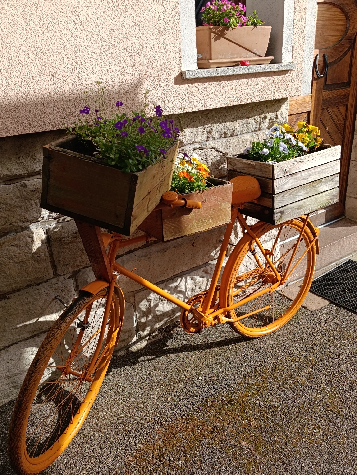 An orange bicycle adorned with wooden planters sits against a light-colored stone building. The front planter contains purple flowers, while the rear planter holds a variety of colorful flowers. A small planter with pink flowers is visible in a window above. The ground is covered in small gravel, and a doormat lies near a wooden door to the right of the bicycle. The location is Unknown, Unknown.