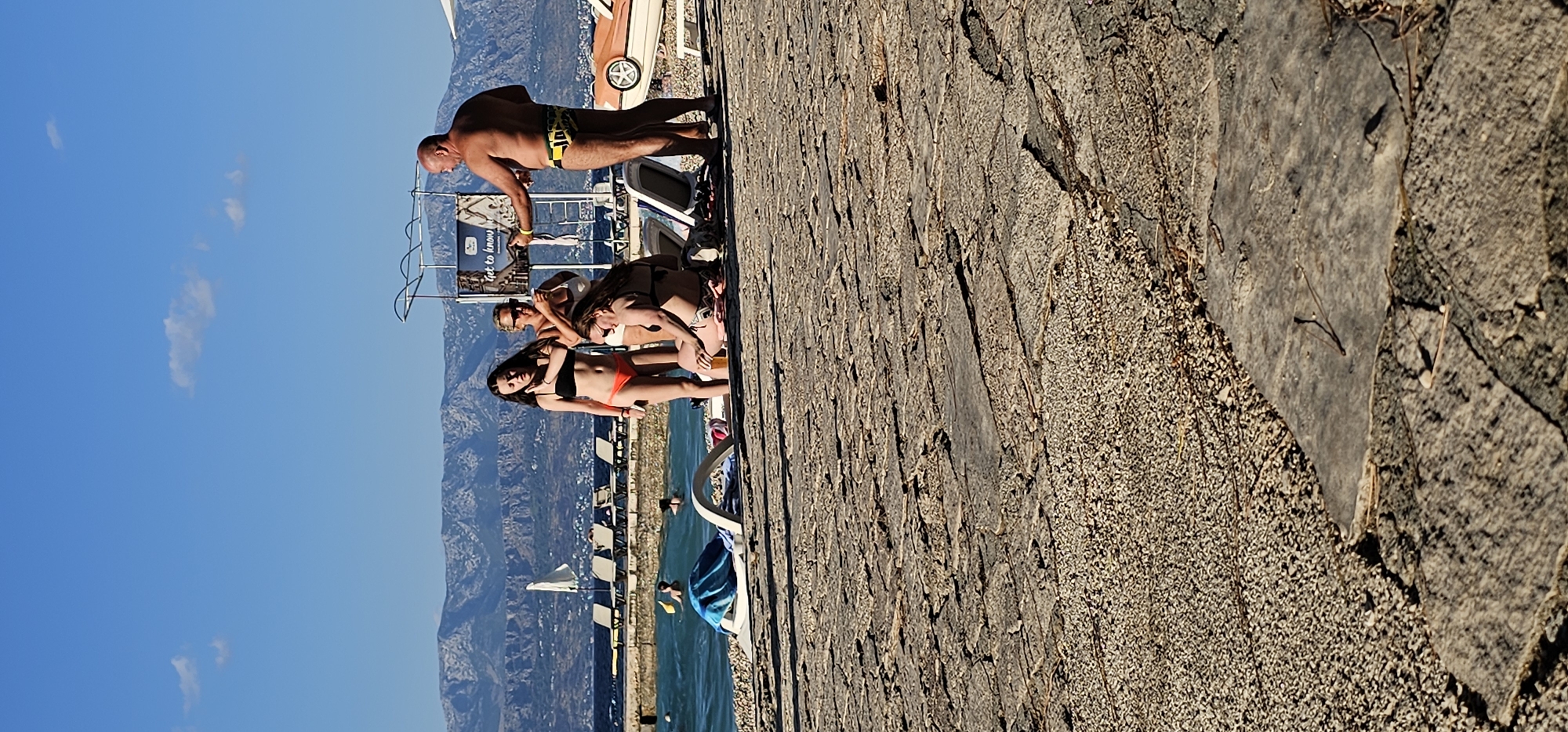 The image shows a sunny day at a beach or waterfront in Grad Supetar, Croatia. The foreground is dominated by a textured, weathered stone or concrete surface, possibly a walkway or pier. In the background, several people are present.
A man in yellow and black patterned swim trunks stands on the stone surface, leaning forward. In front of him, a group of three women are gathered. One woman in a black bikini is sitting and looking towards the camera, while another woman in an orange bikini top and black bottoms is lying down next to her. A third woman, wearing a white bikini, is partially visible behind them. Further back, people are in the water, which appears to be a clear, blue sea. Mountains are visible in the distance, under a bright blue sky with a few scattered white clouds.
A metal railing and a portion of a boat or structure are also visible behind the people on the stone. There is a sign with text on it, but it is largely obscured and difficult to read clearly. The overall impression is of a relaxed, summer day at a popular coastal location.