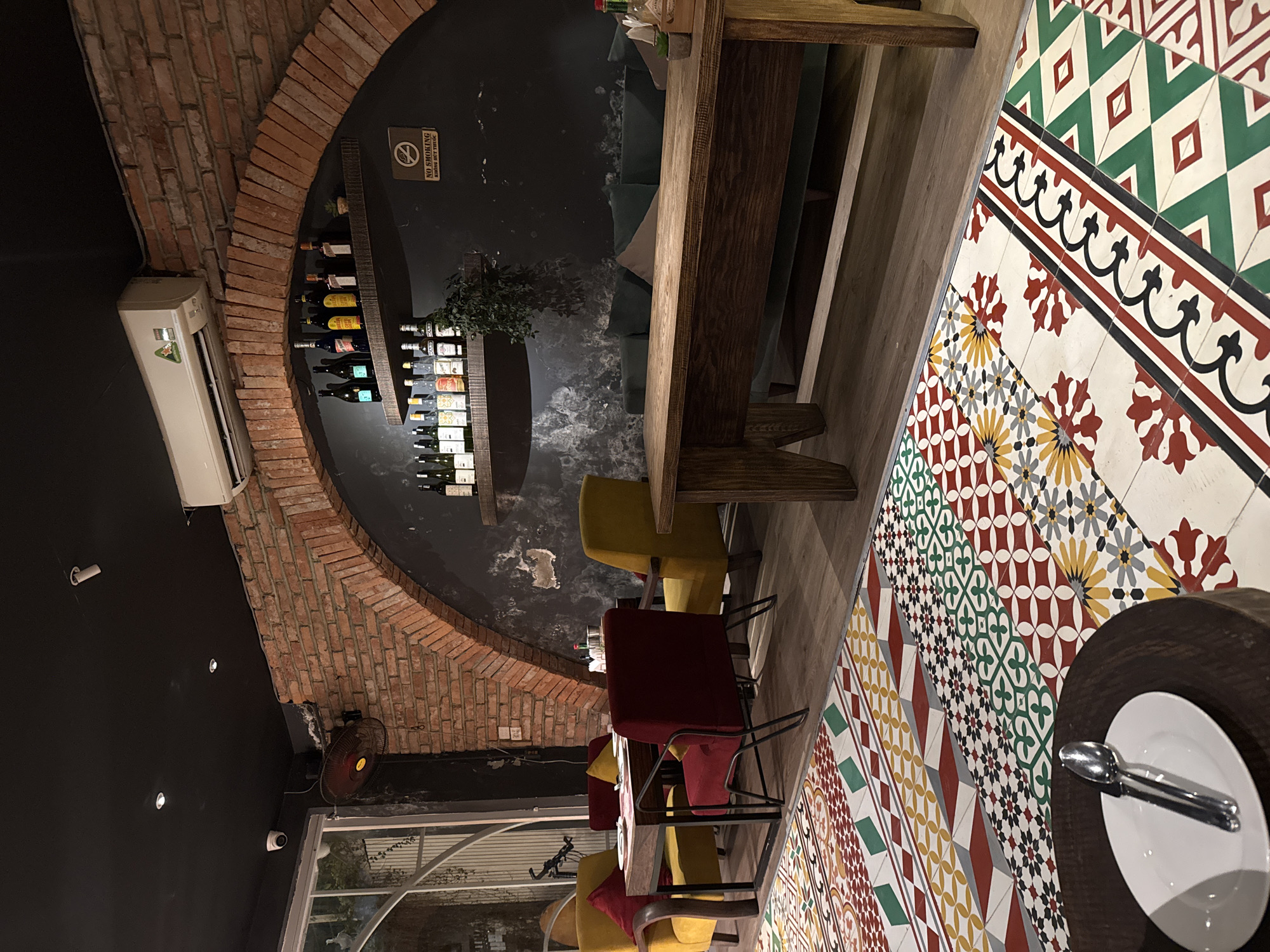 The image depicts the interior of a restaurant in Hanoi, Vietnam. The restaurant features a distinctive tiled floor with intricate geometric patterns in red, yellow, green, and white. A large wooden table sits prominently in the foreground, with a wooden bench and two wooden chairs placed on either side. To the left, a smaller square table is set with plates and red and yellow chairs.
The back wall is constructed of exposed brick, forming a large archway that frames two shelves stocked with various bottles of wine and liquor. A "No Smoking" sign is visible on the wall. Below the shelves, a dark sofa with green cushions is partially visible. A plant hangs from the upper shelf, adding a touch of greenery to the scene. An air conditioning unit is mounted high on the brick wall. The ceiling is painted black and features several recessed lights. A circular security camera is also mounted on the ceiling near the left edge of the image. A red oscillating fan is mounted on the wall near the window on the far left. The overall ambiance suggests a modern yet rustic dining establishment.