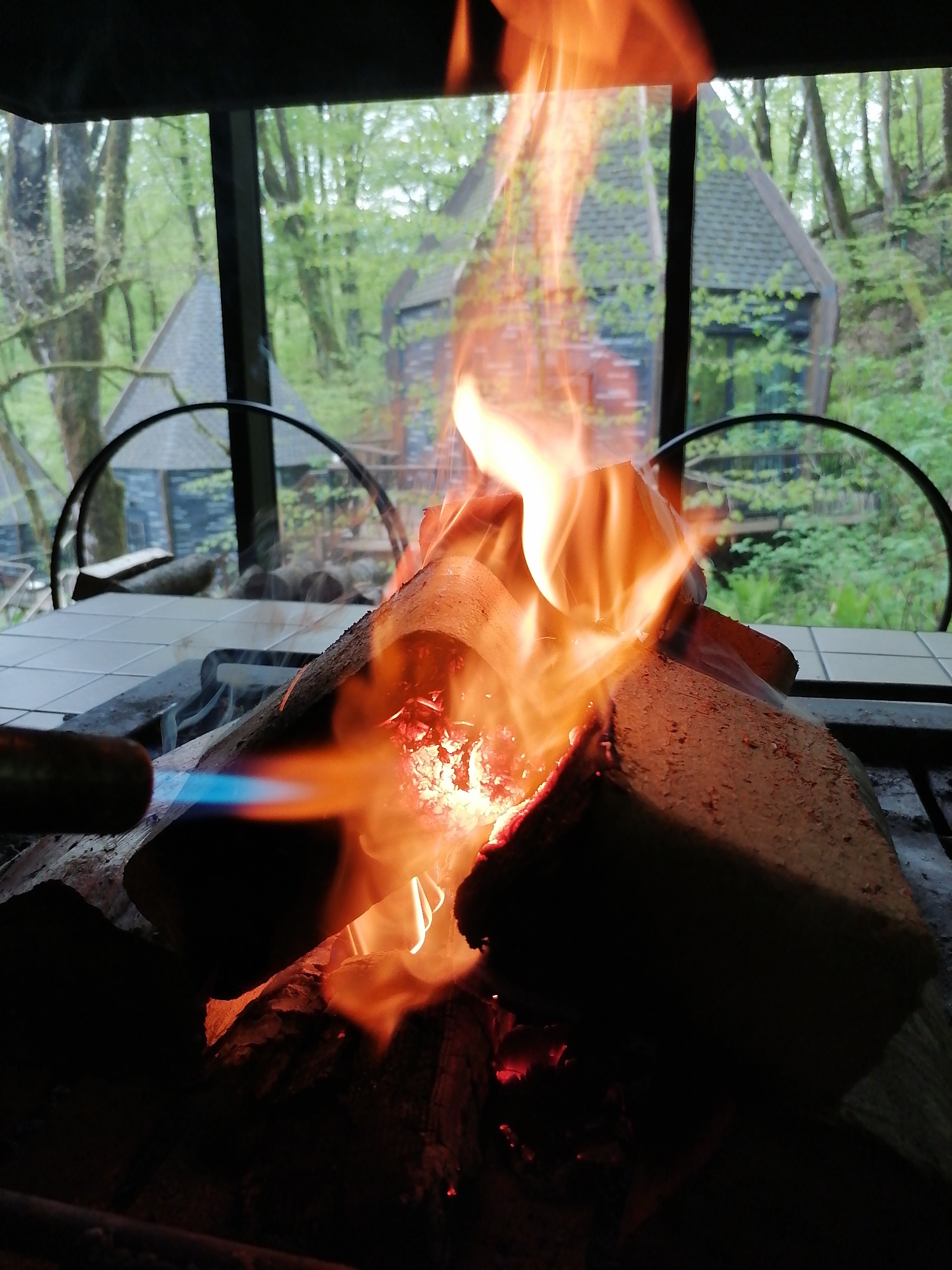 The image captures a close-up view of a fire being ignited or intensified within an indoor or semi-enclosed setting, looking out onto a lush forest scene. In the foreground, a prominent fire burns, with bright orange and yellow flames engulfing several logs. Glowing red embers are visible at the base of the fire. To the left, a blowtorch with a distinct blue flame is being applied directly to the logs, indicating an active process of starting or stoking the fire. The fire appears to be contained within a metal grate, situated on a surface covered with white square tiles.
Through a large window in the background, a verdant forest with tall trees and dense green foliage is visible. The natural light suggests it is daytime, possibly a bright spring or summer day with soft, diffused light, indicating overcast or partly cloudy weather. Nestled among the trees are at least two rustic-looking cabin or lodge-like structures with dark, pointed roofs. These structures have dark walls, and one on the left appears to have faint, indecipherable white writing on its exterior, possibly a sign or blackboard. A black metal ring, perhaps part of a chair or outdoor furniture, is visible between the fire and the window, adding to the outdoor or semi-outdoor aesthetic. The scene suggests a cozy, natural setting, possibly in a recreational area or a home with a strong connection to nature in Medoveevka, Russia.