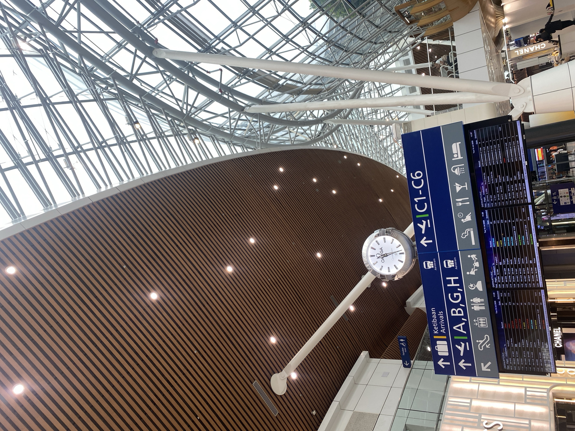The image shows a view from inside Kuala Lumpur International Airport (KLIA) in Sepang, Malaysia. The airport features a modern architectural design with a curved wooden ceiling and a large glass-paneled roof supported by a complex metal structure. A prominent circular clock with Roman numerals and an "OMEGA" logo hangs from a white pole suspended from the ceiling. Below the clock, a large blue departure and arrival information board is visible. The board displays flight information, including destinations, times, and gate numbers. Arrows indicate directions for "Ketibaan Arrivals" and general area access (A, B, G, H). There are also icons representing amenities like food, showers, and luggage. In the foreground, a section of the departure board is clearly readable, showing flight details for July 14, 2026. Further down, a prominent "CHANEL" advertisement is displayed. Two individuals are faintly visible in the background, one seated at a table in a cafe area and another walking near the Chanel advertisement. The lighting suggests it is daytime, with natural light filtering through the glass roof and artificial lights illuminating the interior.