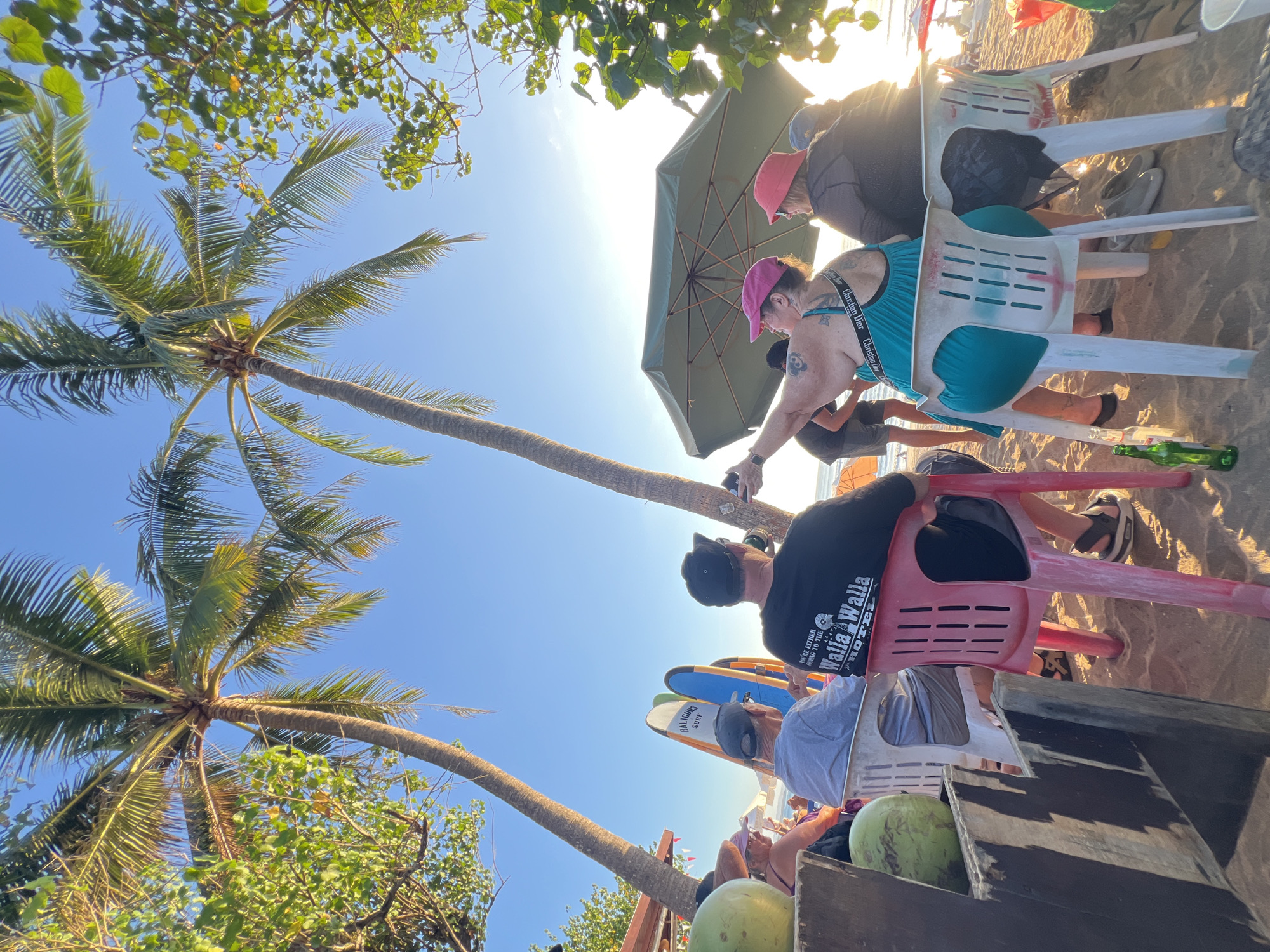 At a beachside establishment in Kuta, Indonesia, several individuals are seated on plastic chairs on the sand, facing away from the camera. Palm trees frame the scene, with their fronds casting shadows. In the background, the ocean is visible, with a clear blue sky above. Surfing equipment, including surfboards, is propped up nearby. One person is holding up a phone, seemingly taking a photo. Another person is wearing a bright pink hat and a teal-colored dress, and appears to be gesturing. A large green umbrella provides shade for some of the seated individuals. Two green coconuts are visible on a wooden surface in the foreground, and a green glass bottle lies on the sand. The overall atmosphere suggests a relaxed beach setting.