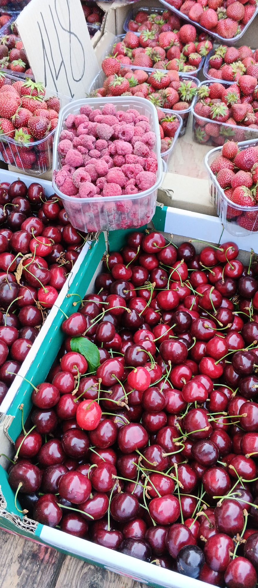 The image shows a close-up of several containers filled with fresh fruit, likely at an outdoor market. In the foreground, a green and white cardboard box is overflowing with dark red cherries, many still attached to their stems. To the left, another similar box also contains cherries. In the upper portion of the image, a clear plastic container is filled with bright pink raspberries. Behind the raspberries, several plastic containers are visible, filled with vibrant red strawberries. A partially visible cardboard sign with handwritten markings is positioned in the upper left quadrant. The overall impression is one of abundance and freshness, with the fruits neatly arranged in their respective containers. The scene is likely a fruit stall at a market in Smila, Ukraine.