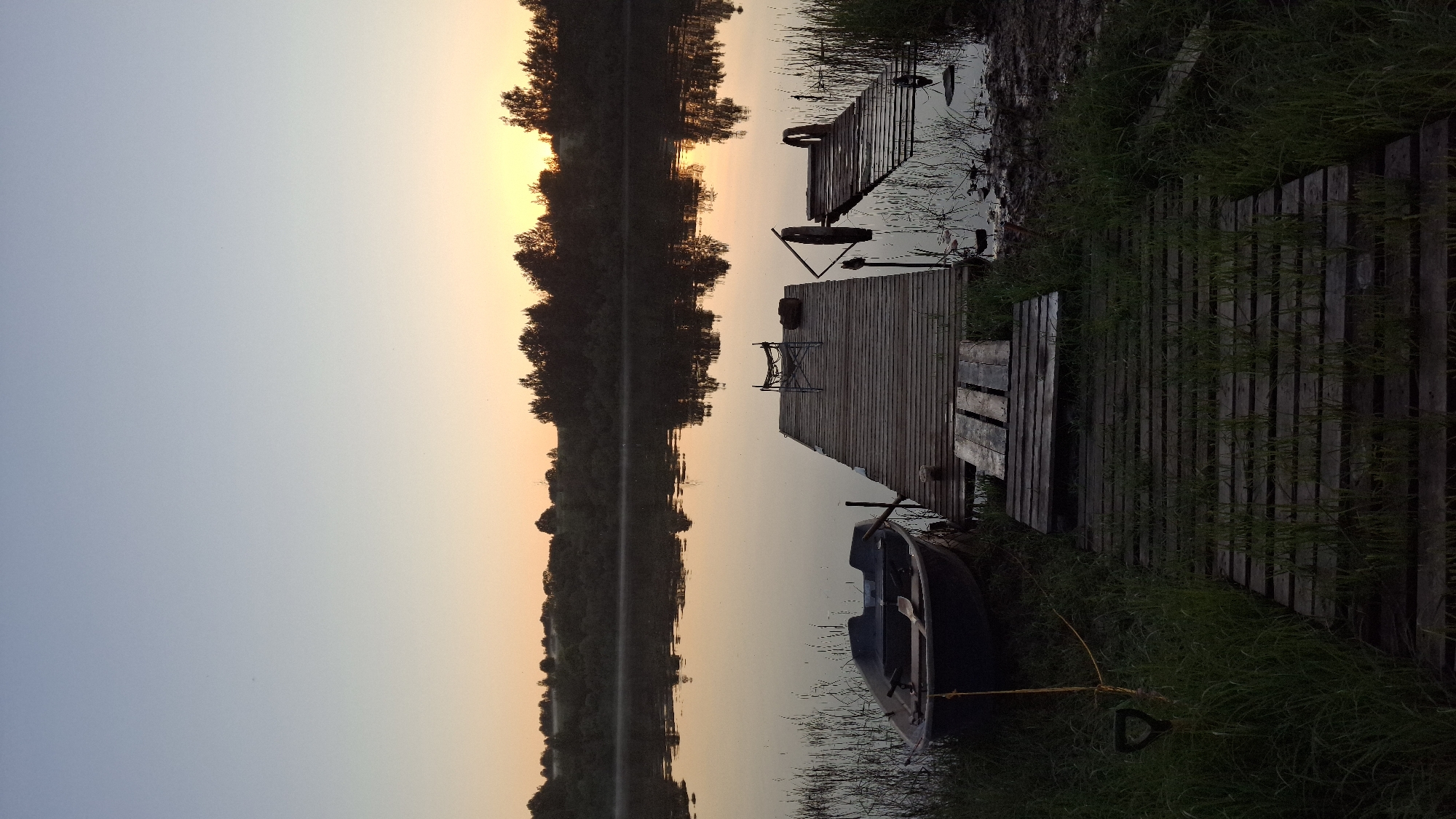 This forensic description details an outdoor scene in Sitno, Russia, depicting a tranquil waterfront at either sunrise or sunset. The upper half of the frame shows a clear sky transitioning from a pale blue at the zenith to a warm orange-yellow glow on the distant horizon. A dense line of dark green trees and foliage forms the opposing bank of a calm body of water, their silhouettes reflected on the water's surface.
In the midground, the water's surface is smooth and reflective, mirroring the soft sky colors and distant vegetation. In the foreground, on the left, a dark-colored rowboat is partially beached on the grassy bank, with its stern slightly in the water. A yellow rope extends from the boat to a dark, D-shaped metal object embedded in the grass. Tall, green grass covers the immediate shoreline and grows between the wooden planks of a pathway that extends from the bank towards the water. This pathway leads to a rectangular wooden pier. A light-colored, collapsible chair is positioned on the pier, near a dark, cylindrical object. To the right of the main pier, a partially submerged and broken section of a wooden dock is visible, along with a dark, circular object, possibly a tire, also partially submerged. The immediate shoreline near the docks shows areas of exposed mud and sparse reeds growing from the water on the far right. No individuals are present in the scene.
