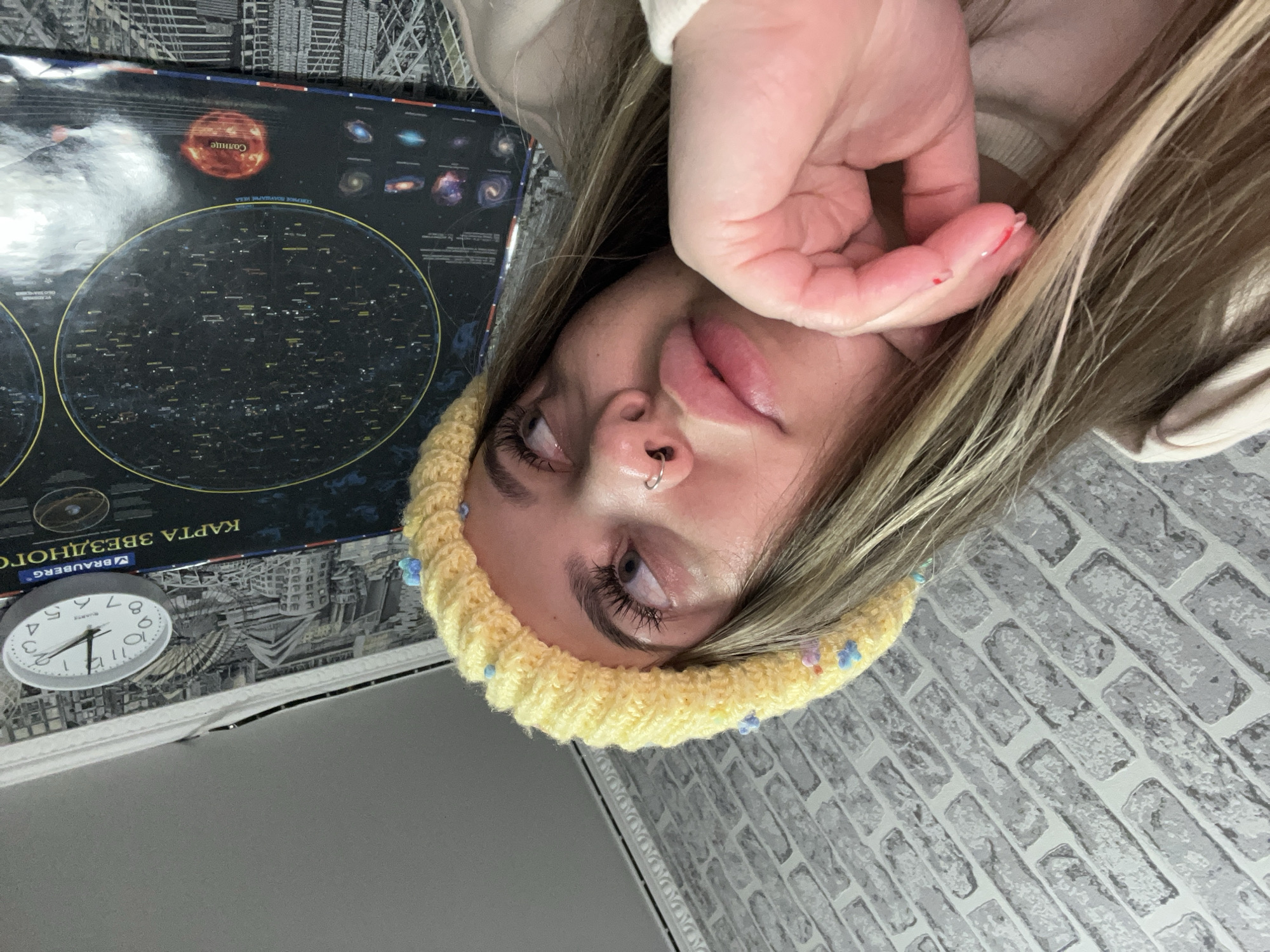 A selfie-style image shows a person with light blonde hair, fair skin, and a nose ring, wearing a yellow knitted hat with small blue and purple decorations. The person's face is tilted, with their hand partially covering their face. The thumb has a small red mark on the nail.
To the left of the person, there's a star chart with a depiction of the sun and constellations. A round clock with hands indicating approximately 11:58 is also visible. The background consists of a light gray brick-patterned wall and a patterned material with images of city buildings.
There is no information in this image to determine if it was taken in Novopokrovskaya, Russia.