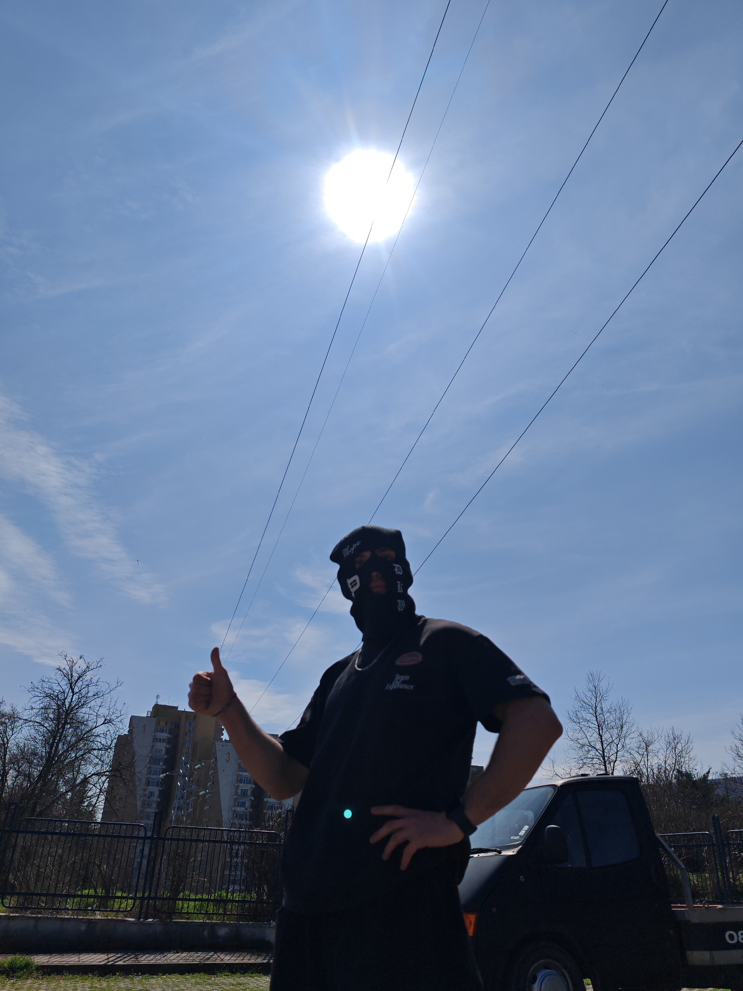 A daylight outdoor photograph captures a male subject standing in a residential area of Shumen, Bulgaria, characterized by post-socialist era apartment blocks. The subject, positioned slightly right of center, faces forward and presents a thumbs-up gesture with his right hand. He is dressed in all black, consisting of a balaclava obscuring his face and head, black sunglasses, a black beret with white text "Olype", and a black short-sleeved polo shirt featuring a white "Begin Experience" logo on the left chest. A silver-colored chain is visible around his neck, and a black wrist-worn device is on his left wrist. His left hand rests on his hip.
To the subject's right and slightly behind, the front section of a dark-colored (likely black) pickup truck or small flatbed truck is partially visible, parked on a paved surface. The numbers "08" are discernible on the side or rear of the truck's bed. Further back and to the left, behind a dark metal fence and a line of bare deciduous trees, a multi-story concrete apartment building with multiple balconies stands prominently. The building's facade shows the numbers "1" and "3".
The sky is bright blue with scattered thin white clouds. The sun, a strong source of light and glare, is located in the upper left quadrant of the frame, causing a lens flare. Several dark, thin utility lines stretch diagonally across the upper sky. The immediate foreground shows a paved surface with some visible grass.