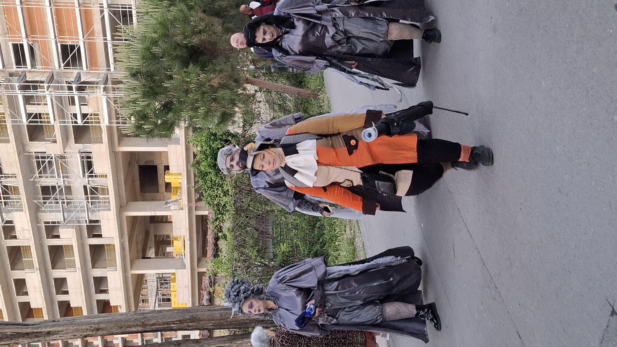 The image shows three people in costume on a sidewalk in an urban setting. One person in the center is wearing an orange, white, and tan patchwork costume with black pants and shoes. The other two are wearing grey, shiny, full-length costumes. A construction site is in the background on the left with a building under construction. There are trees and greenery between the construction site and the people. The sidewalk is grey concrete.