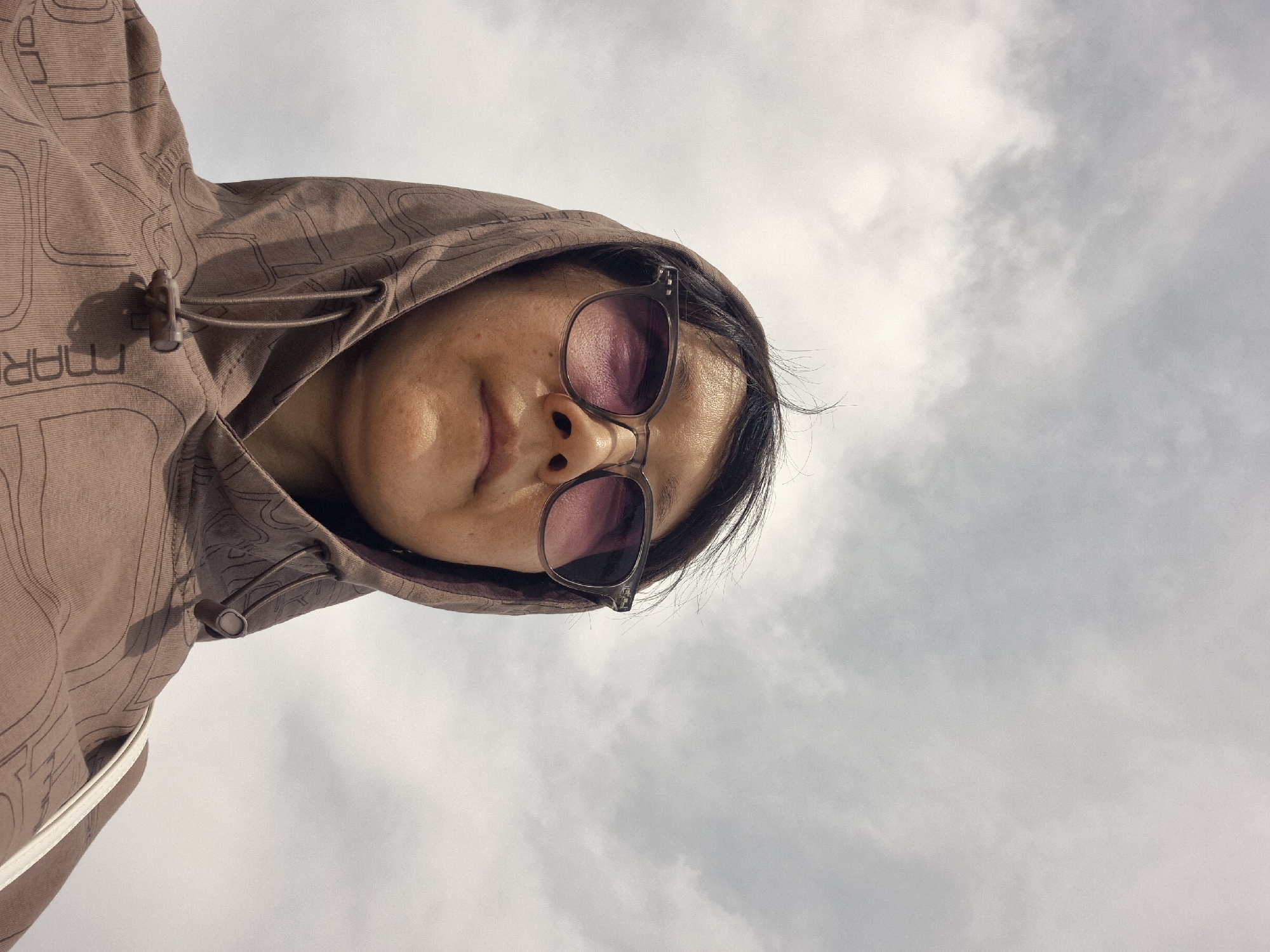 A low-angle, waist-up shot shows a person wearing sunglasses and a brown hooded sweatshirt. The person's face is partially obscured by the hood and sunglasses. The background is a cloudy sky. The image does not provide any information about the location in Kokshetau, Kazakhstan.
