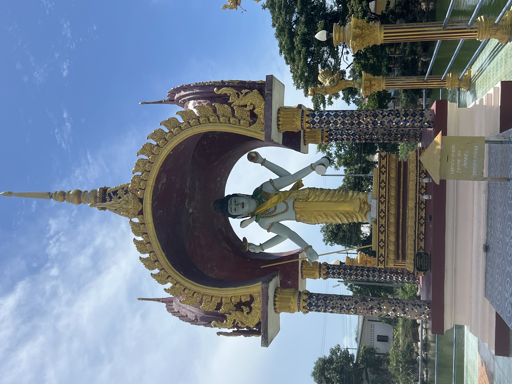 The image shows a statue of a figure with multiple arms in Baan Plai Laem, Thailand. The statue is gold and has a headdress. It is positioned inside a large ornate arch, with a red interior and gold decorative trim, that is supported by decorated columns. The sky is visible behind the statue, which appears to be in a garden area with trees and a pond or canal in the background. A donation box is placed to the right of the statue.