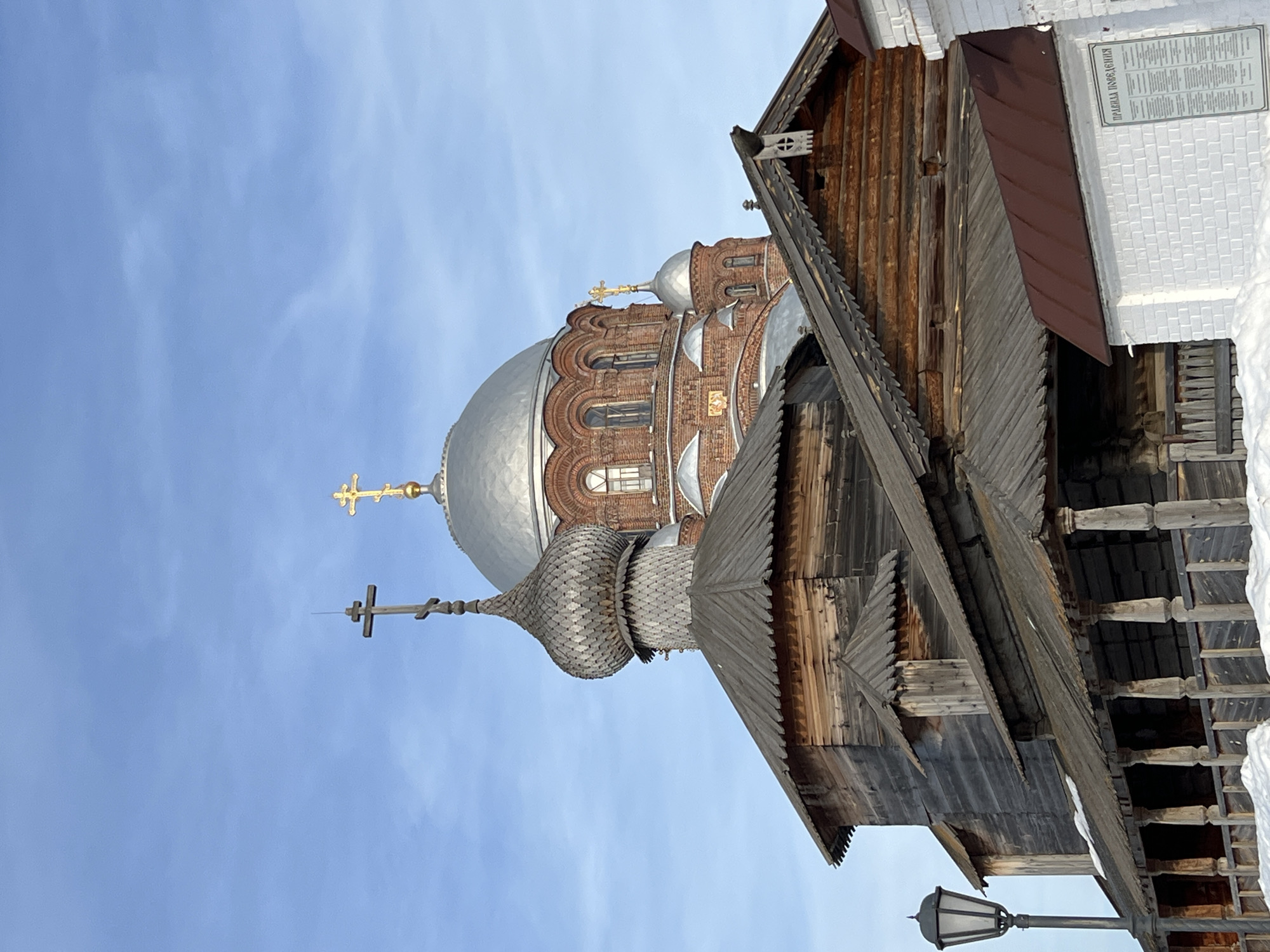 The image displays a winter scene in the city of Sviyazhsk, Russia, featuring two prominent Orthodox church structures under a clear, light blue sky with minimal cirrus clouds.
In the foreground and midground, a dark, weathered wooden church is visible. Its roof is composed of dark, flat shingles, and a distinct onion-shaped dome, constructed from overlapping wooden scales or shingles, rises from it. This dome is surmounted by a simple, unadorned wooden Orthodox cross. The visible walls of this structure consist of horizontal, dark-stained or aged timber logs. To the left, an antique-style black street lamp on a pole stands adjacent to the wooden church. Portions of a wooden veranda or walkway with turned posts and a railing are discernible at the base of the wooden structure.
Behind and partially above the wooden church, a larger, reddish-brown brick cathedral dominates the background. It features a large, smooth, silver-colored main dome, capped with a golden Orthodox cross. Additional smaller silver domes, also adorned with golden Orthodox crosses, are present on the brick structure, which exhibits arched windows and ornamental brick detailing.
To the lower right, a section of a white brick wall topped with a dark red metal roof is visible. A rectangular sign with Cyrillic text, reading "ПРАВИЛА ПОВЕДЕНИЯ" (Rules of Conduct), is attached to this wall. Accumulations of snow are present on the ground and on lower roof sections, indicating a cold environment. No individuals are present in the depicted scene.