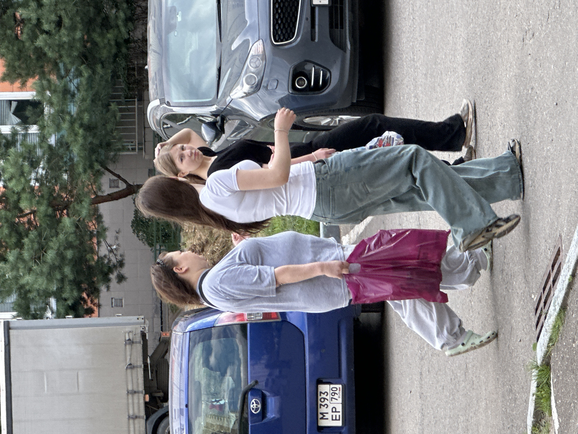 Three individuals are walking on a paved area in Podolsk, Russia, near parked vehicles and trees.
In the foreground, a person wearing a light gray t-shirt and light gray sweatpants walks from left to right, carrying a bright pink plastic bag in their right hand. They are wearing light green clogs. To their right, a second person, wearing a white t-shirt and loose-fitting teal jeans, also walks from left to right, their left hand held out slightly. They are wearing dark shoes. Behind these two individuals, a third person in a black t-shirt and black pants walks from left to right, their right hand resting on their head, and their left hand is obscured. They are wearing dark-colored athletic shoes.
To the left of the pedestrians, a blue hatchback car is parked, with its rear visible. A license plate reading "M 393 EP 790 RUS" is affixed to the rear. To the right of the pedestrians, a dark grey SUV is parked. A large, silver truck trailer is visible in the background on the far left. Trees with green foliage are present behind the parked vehicles, and a building with windows is partially visible in the background. The ground is paved with asphalt, with some grass and a metal grate visible near the bottom left of the frame.