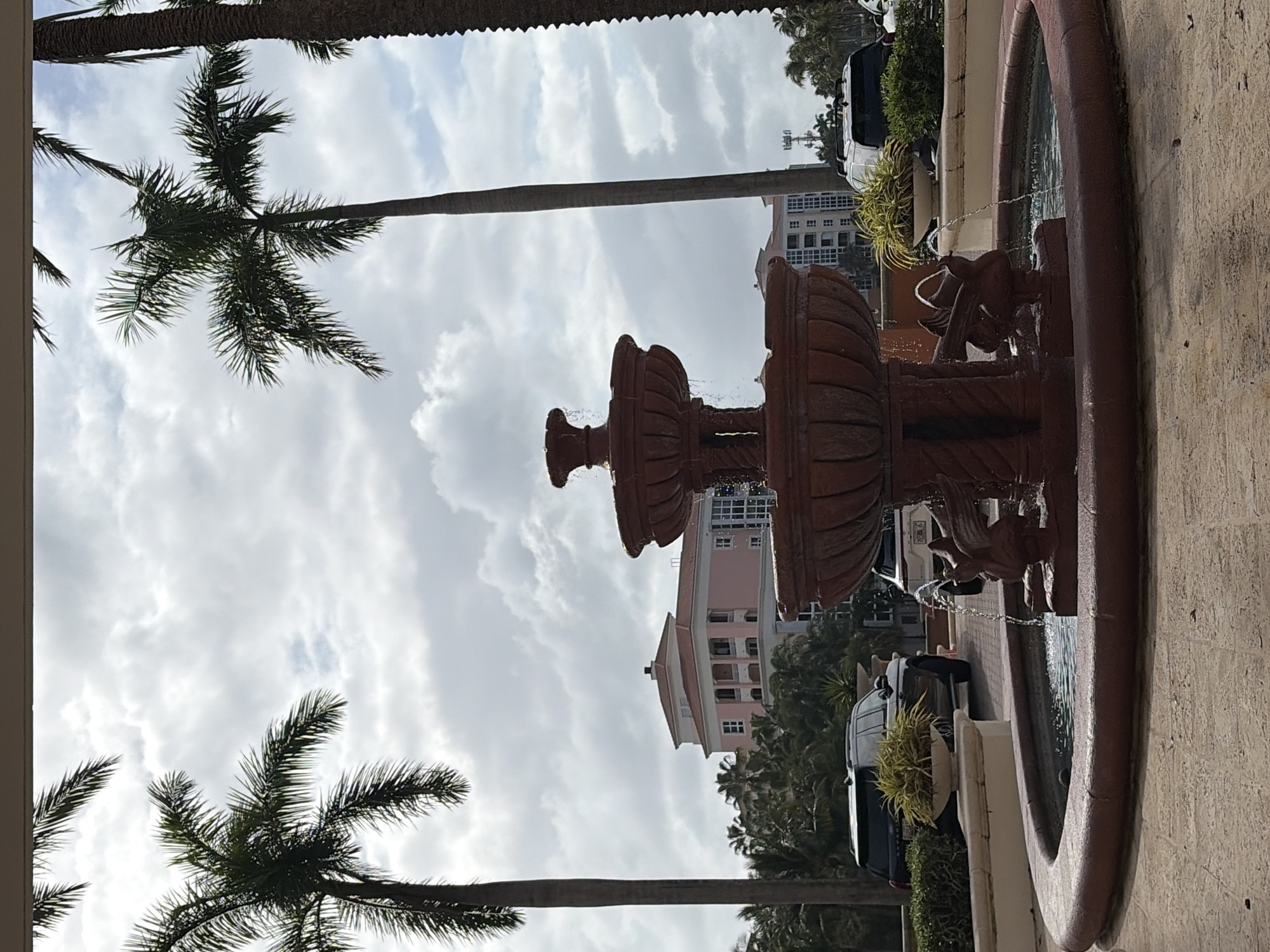 A ground-level, medium-shot photograph features a decorative terra-cotta fountain in the foreground with a Key Biscayne, Florida, United States background. The fountain is multi-tiered, with water flowing from spouts shaped like animal heads into a basin. The basin is bordered by a textured, slightly curved wall. Beyond the fountain, palm trees are visible against a cloudy sky. Several buildings, including a pink building with multi-story levels and a building with numerous windows, are partially visible between the palm trees. Several cars and potted plants, parked along a street are also in the view.