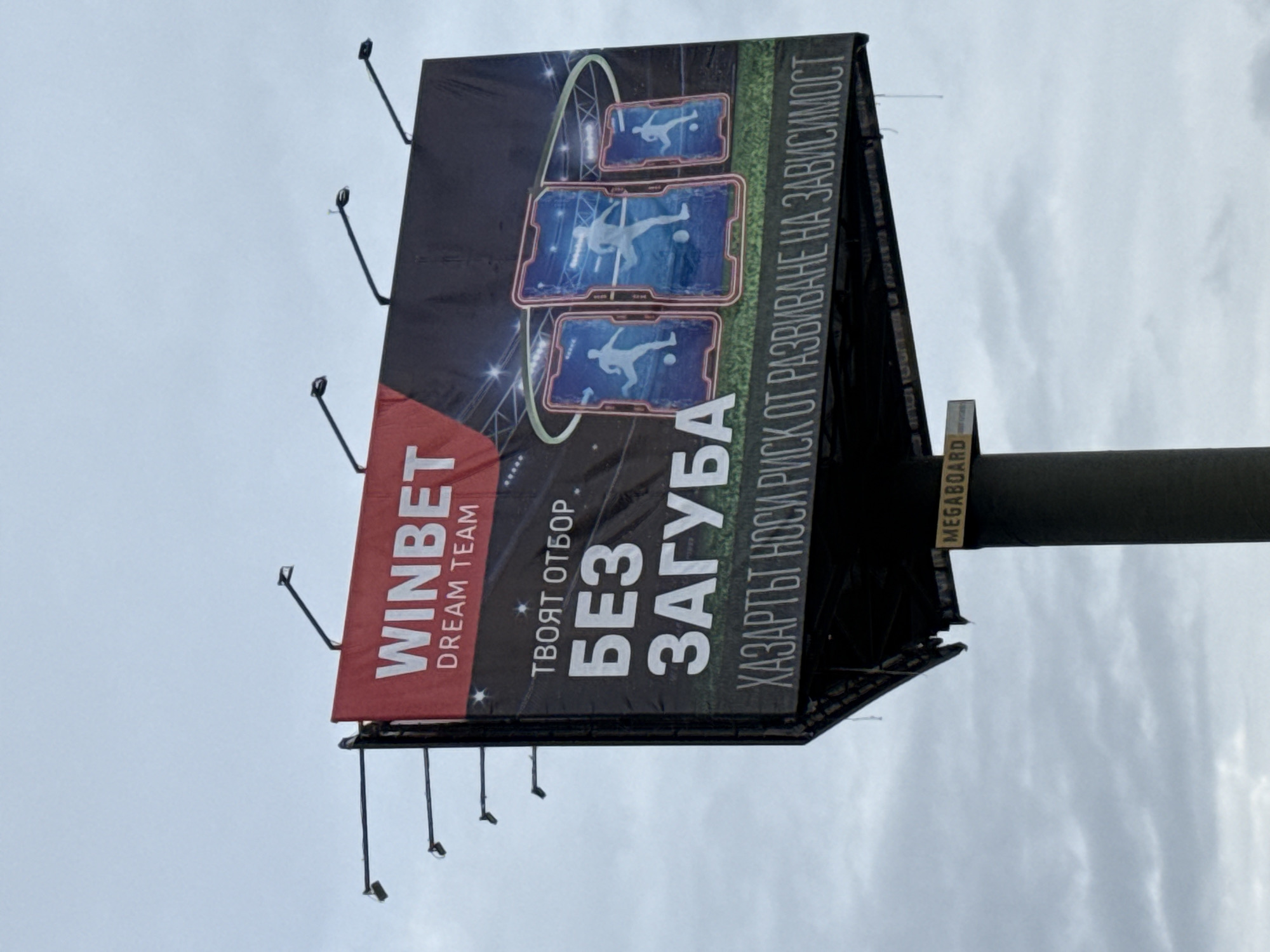 The image is a billboard featuring an advertisement for WINBET, a betting company, situated in Sofia, Bulgaria. The sign displays the company logo, the phrase "Dream Team", and the slogan "ТВОЯТ ОТБОР БЕЗ ЗАГУБА" (Your Team Without Losses). It includes imagery of a soccer player in a digital, futuristic format. There is also a warning message: "ХАЗАРТЪТ НОСИ РИСК ОТ РАЗВИВАНЕ НА ЗАВИСИМОСТ" (Gambling carries the risk of developing an addiction). The billboard structure is visible with supporting poles and the word "MEGABOARD" is printed on a portion of the structure. The sky is overcast.