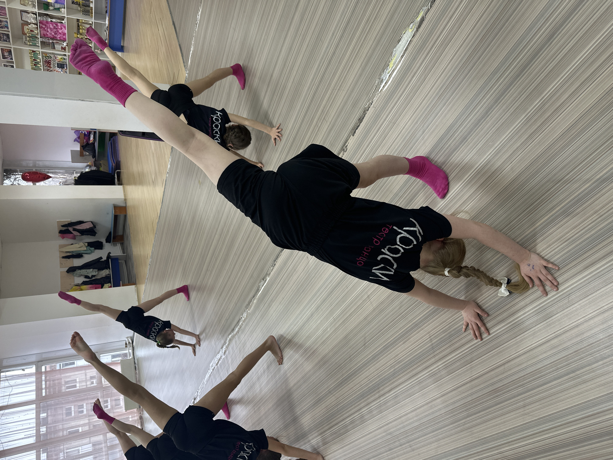 The image depicts a group of young girls performing a stretching or gymnastic exercise in a dance or acrobatics studio located in Votkinsk, Russia. The scene is captured from an angled perspective, looking down at the participants.
At least four girls are visible, all dressed in matching black t-shirts, black shorts, and bright pink socks. They are positioned with their hands on the light-colored, striped wooden floor, extending their legs upwards and outwards in a preparatory pose for a handstand or similar acrobatic movement. The white Cyrillic text on their t-shirts clearly reads "КРАСКИ" (KRASKI, meaning "Paints" or "Colors") and "ТЕАТР ТАНЦА" (TEATR TANCA, meaning "Theater of Dance"), identifying their group or studio. One girl in the foreground has a blonde braid secured with a white hair tie and bow.
The studio environment includes white walls, large mirrors reflecting parts of the room, and a light-colored wooden or laminate floor. In the background, shelves are laden with numerous trophies, awards, and various other items, signifying a place of achievement. A window provides natural light and offers a glimpse of an exterior building. Other objects such as blue mats and a red balloon are also present in the background. The overall atmosphere suggests an active and focused training session for dance or performance arts.