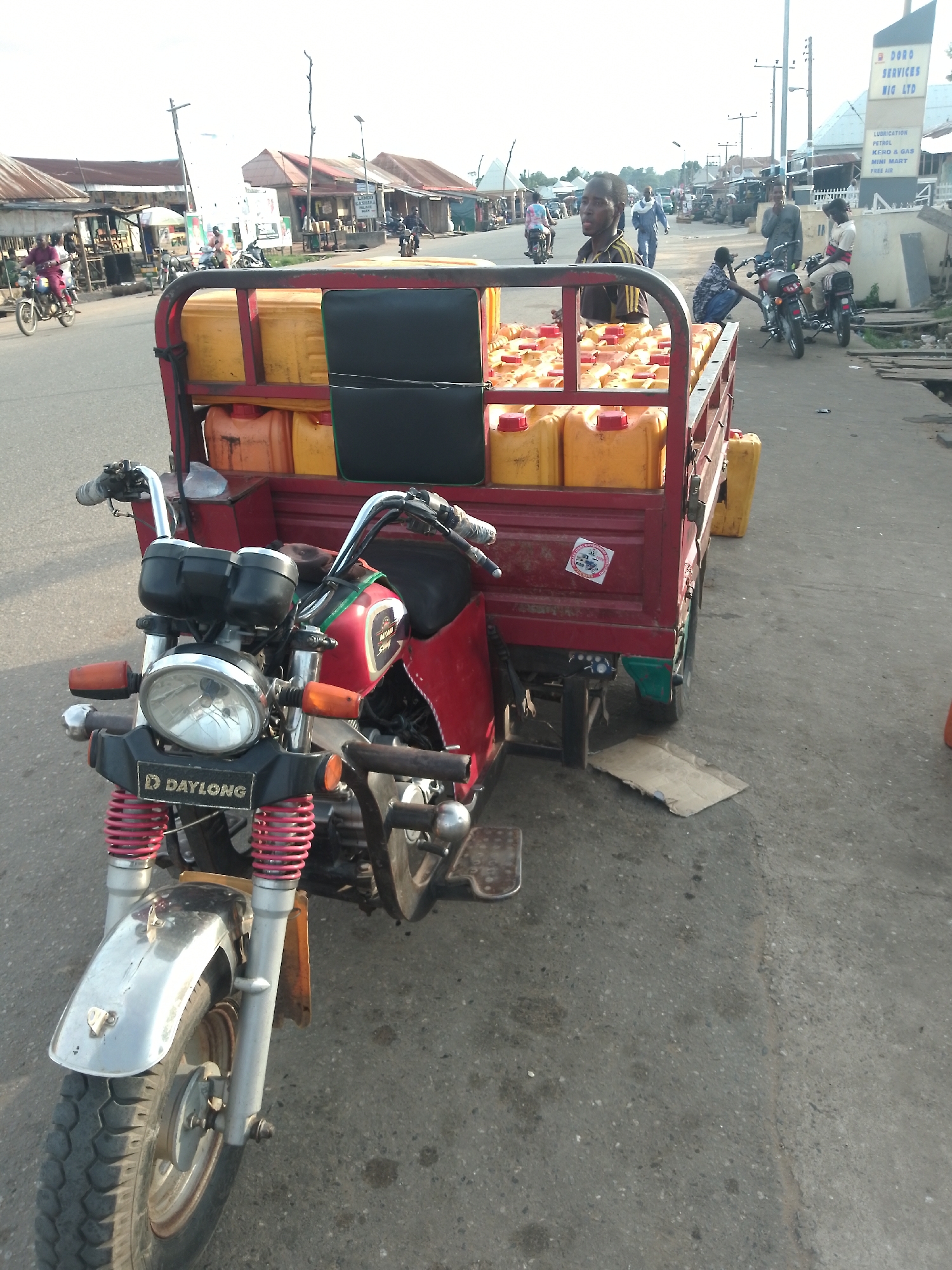 This image depicts a street scene in Serti, Nigeria, likely in the morning or late afternoon given the warm, diffused sunlight. The central focus is a red motorized tricycle loaded with numerous yellow plastic jerrycans, likely containing fuel or other liquids for sale. The tricycle is parked on a paved road, with a person's head visible leaning over the load, suggesting they are the driver or an attendant.
In the background, a row of buildings with corrugated metal roofs lines the street, indicative of local shops and businesses. A prominent sign is visible on the right, with text that reads "DORO SERVICES NIG LTD," followed by "LUBRICATION, PETROL, KERO & GAS, MINI MART, FREE AIR." This further reinforces the commercial nature of the setting.
Several other individuals are present in the scene. A person is riding a motorcycle in the mid-ground, and another individual is kneeling near a parked motorcycle, possibly engaged in conversation or some form of transaction. More people can be seen further down the street, suggesting a bustling, active marketplace or transport hub. The overall atmosphere appears to be one of daily commerce and activity, typical of a roadside market in a Nigerian town. The condition of the road and the buildings suggests a somewhat rural or semi-urban environment.