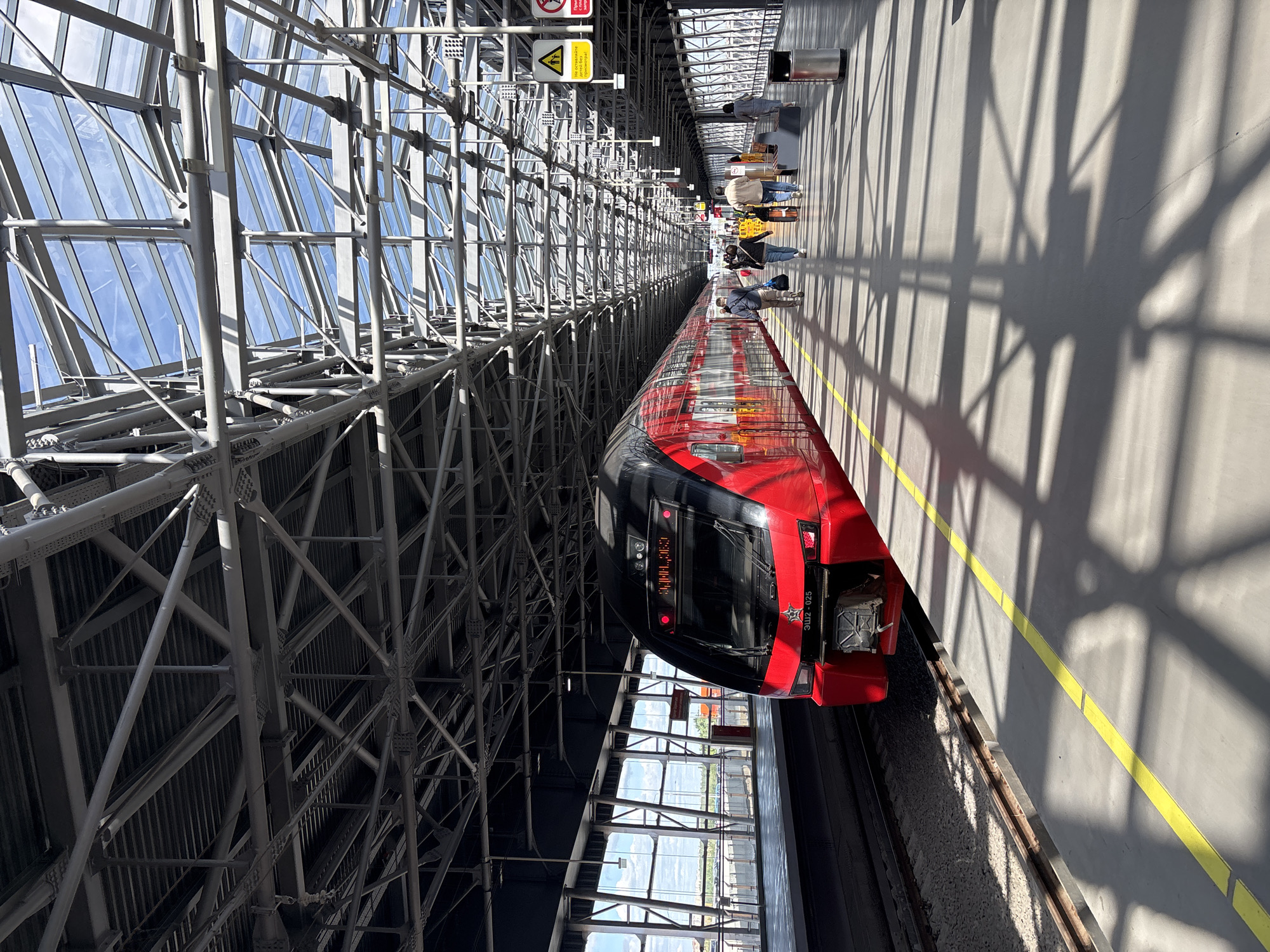An elevated, side-angled perspective captures a red train at a station in Khimki, Russia. The train is parked parallel to a platform where several individuals are waiting. A complex network of steel beams supports the roof of the station, casting shadows on the platform. Overhead signage is visible, and light reflects off the station’s glass roof.