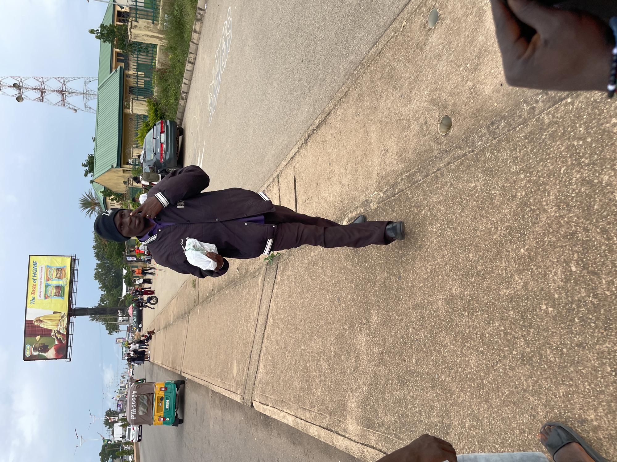 A man in a black beanie and a dark jacket is walking on a concrete sidewalk next to a street. He is holding a white plastic bag in his right hand and has his left hand raised, with his index finger pointing towards his forehead. In the background, there are vehicles, including a colorful tricycle, and a billboard advertising "The Taste of HOME." A telecommunications tower is visible in the distance, along with buildings and trees. The scene appears to be in Jos, Nigeria, based on the context provided. The sky is overcast, indicating it might be evening or a cloudy day.