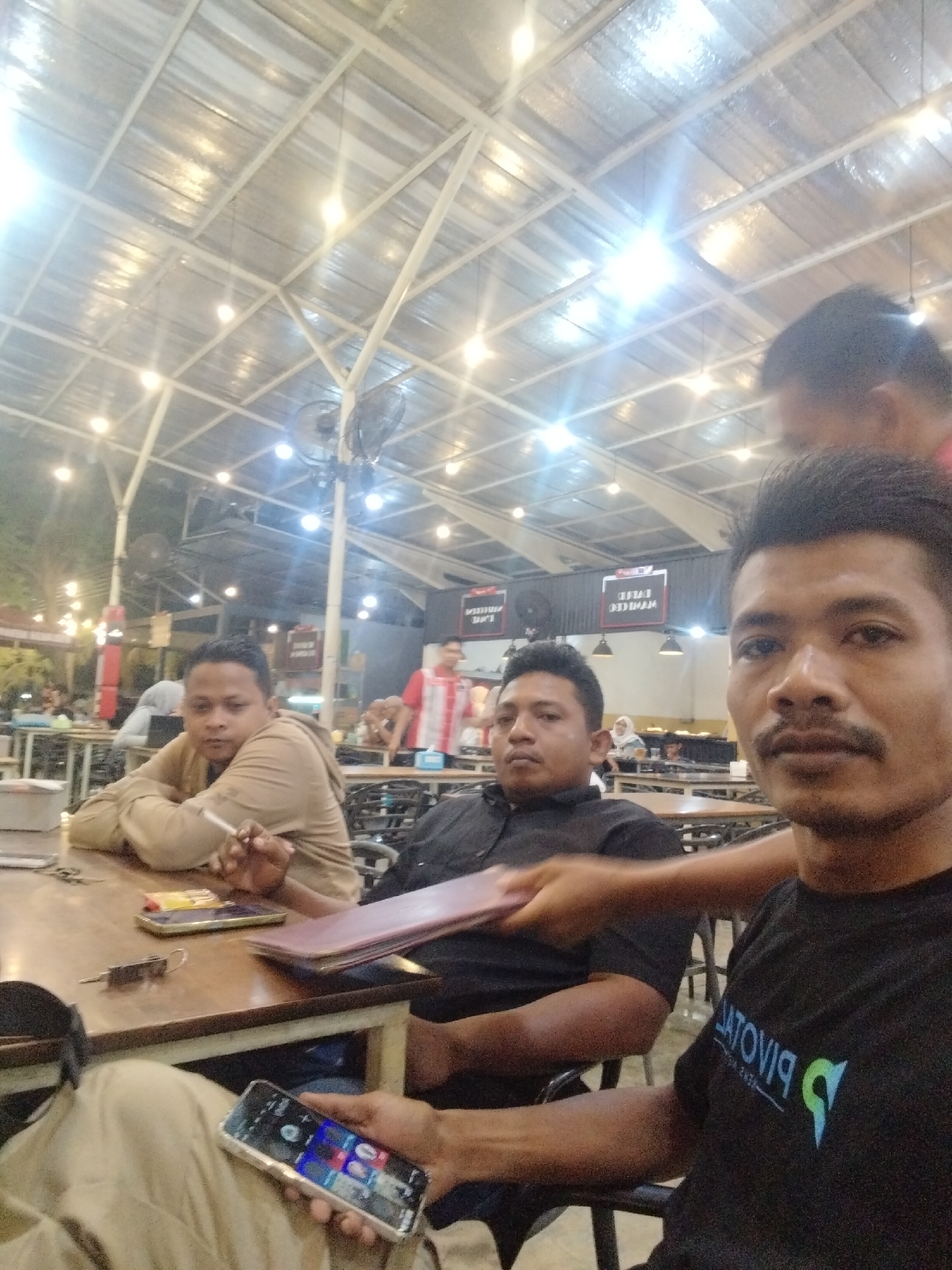 A medium, angled indoor shot depicts three men at a table in what appears to be a restaurant in Lhokseumawe, Indonesia. The man in the foreground is holding a smartphone and wearing a black t-shirt with a logo. Behind him to the left is a man in a black collared shirt. Another man is next to him, wearing a beige jacket and holding a cigarette. A hand extends into the frame, apparently serving or passing a menu to the man in the black shirt. The table has personal items on it, including keys and other phones. The restaurant is populated with other people. The ceiling has a metal frame structure and visible lighting fixtures.