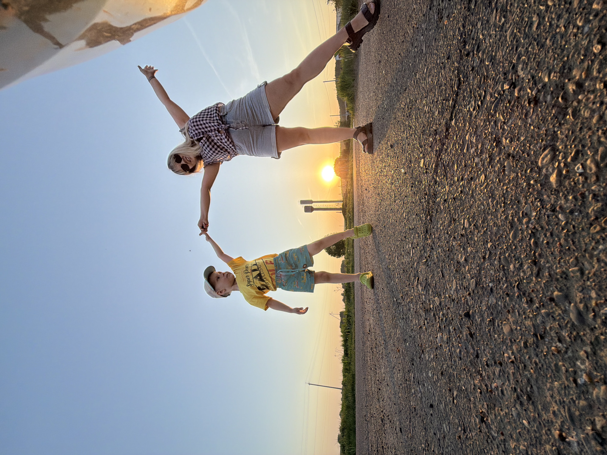 A woman and a child are posing for a picture. The picture has been rotated to make it look like they are suspended in the air. The woman is wearing jean shorts, a checkered top, sunglasses, and sandals. The child is wearing a yellow t-shirt, patterned shorts, a hat, and green shoes. They are holding hands. They are standing on an asphalt road, with a field and power lines in the background. The sun is setting in the background. It is not possible to confirm the location.