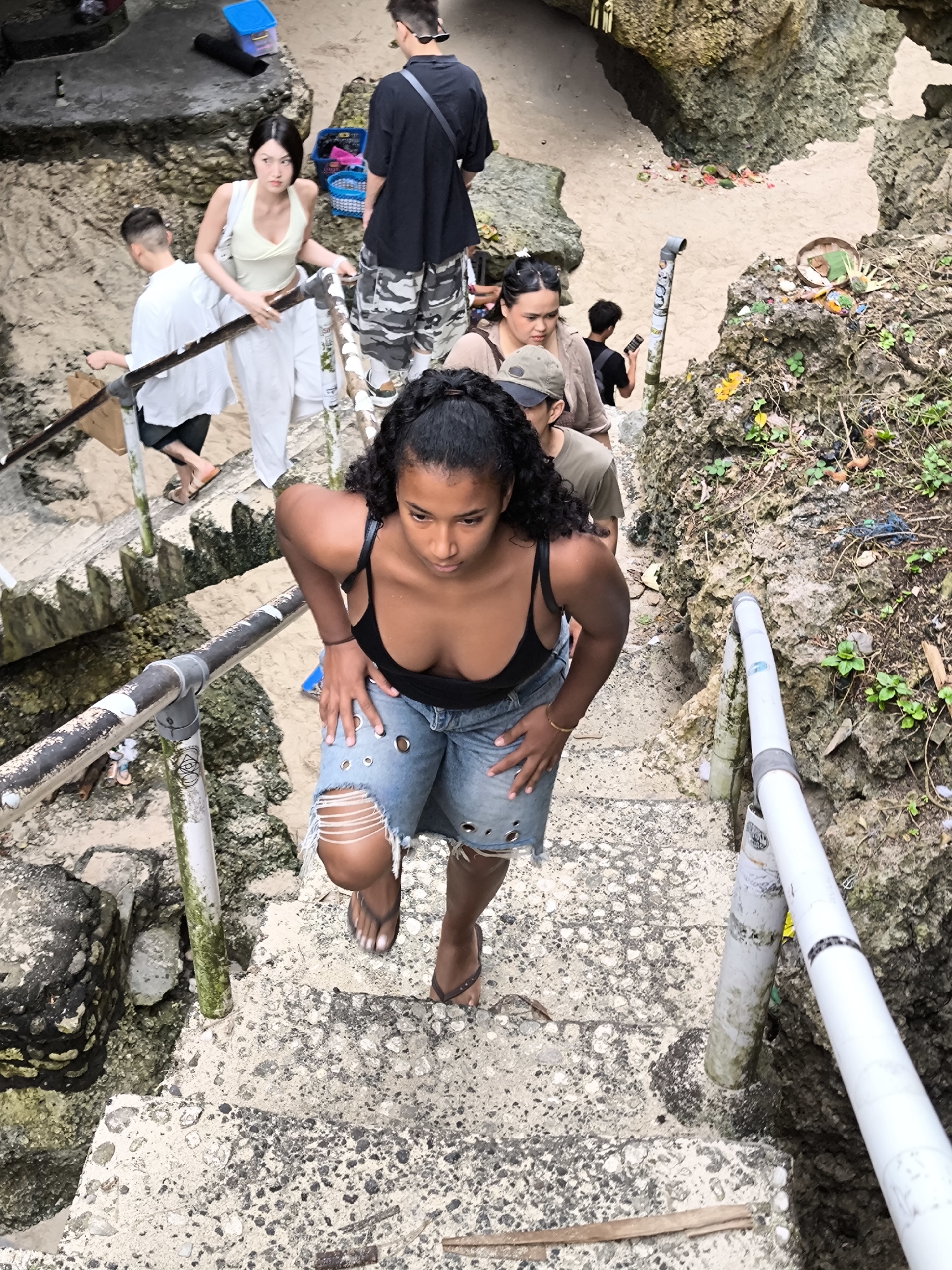 The photograph, taken at Uluwatu, Indonesia, depicts a group of people navigating a weathered concrete staircase. In the foreground, a woman with dark curly hair and a black tank top is ascending the stairs, wearing distressed denim shorts and flip-flops. Behind her, a small cluster of individuals are also making their way up the steps. To the left and right of the staircase, metal railings run along the sides, some sections showing signs of age and discoloration. The surrounding environment is characterized by rocky terrain and some vegetation. In the background, a beach and rocky cliffs can be seen.