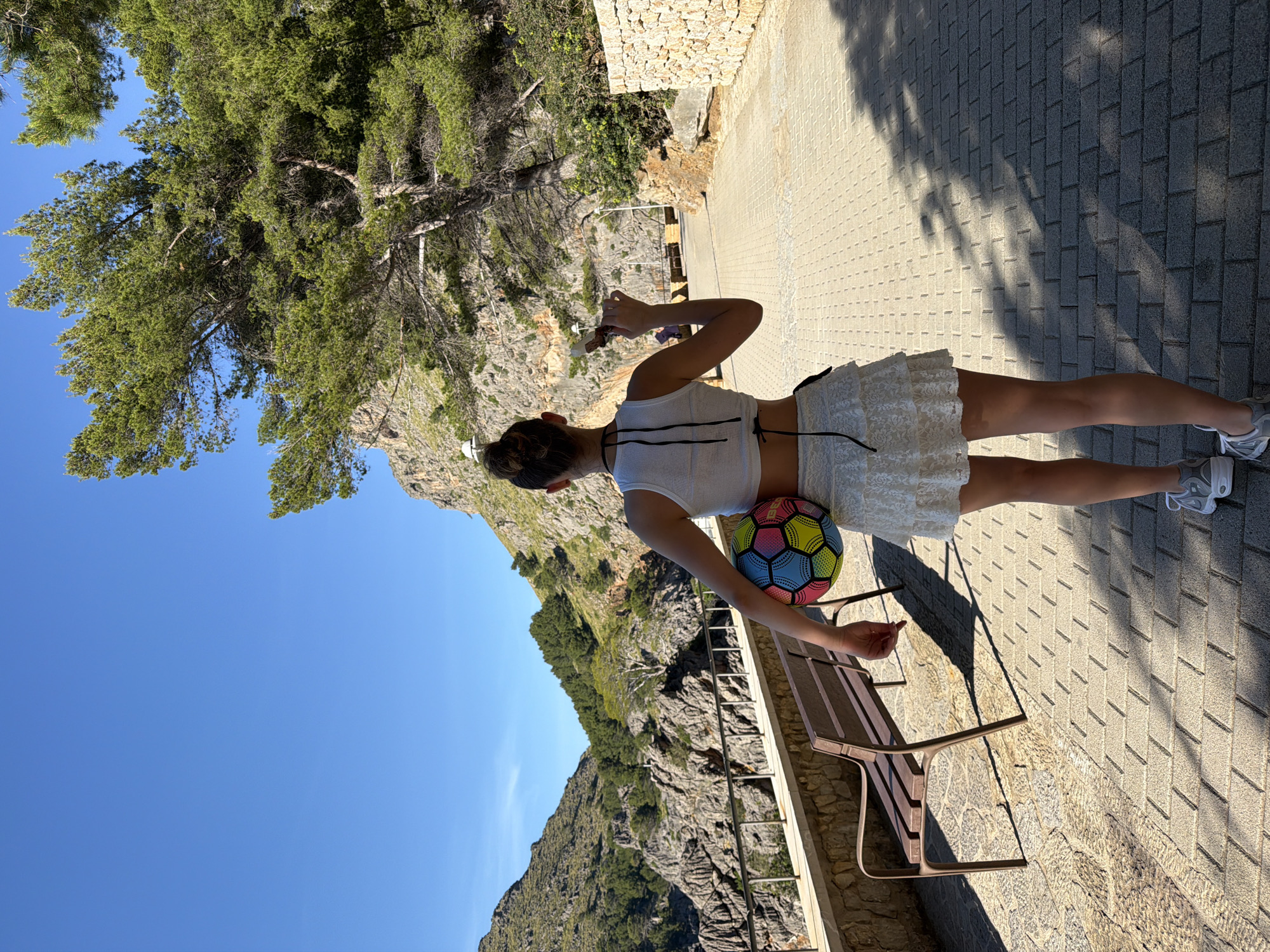 A young woman stands on a stone-paved walkway next to a metal bench, with a colorful ball tucked under her arm. She faces away from the viewer, looking towards a mountainous landscape in Escorca, Spain. A large tree looms in the background, partially obscuring a clear blue sky. The walkway and bench are partly shaded.