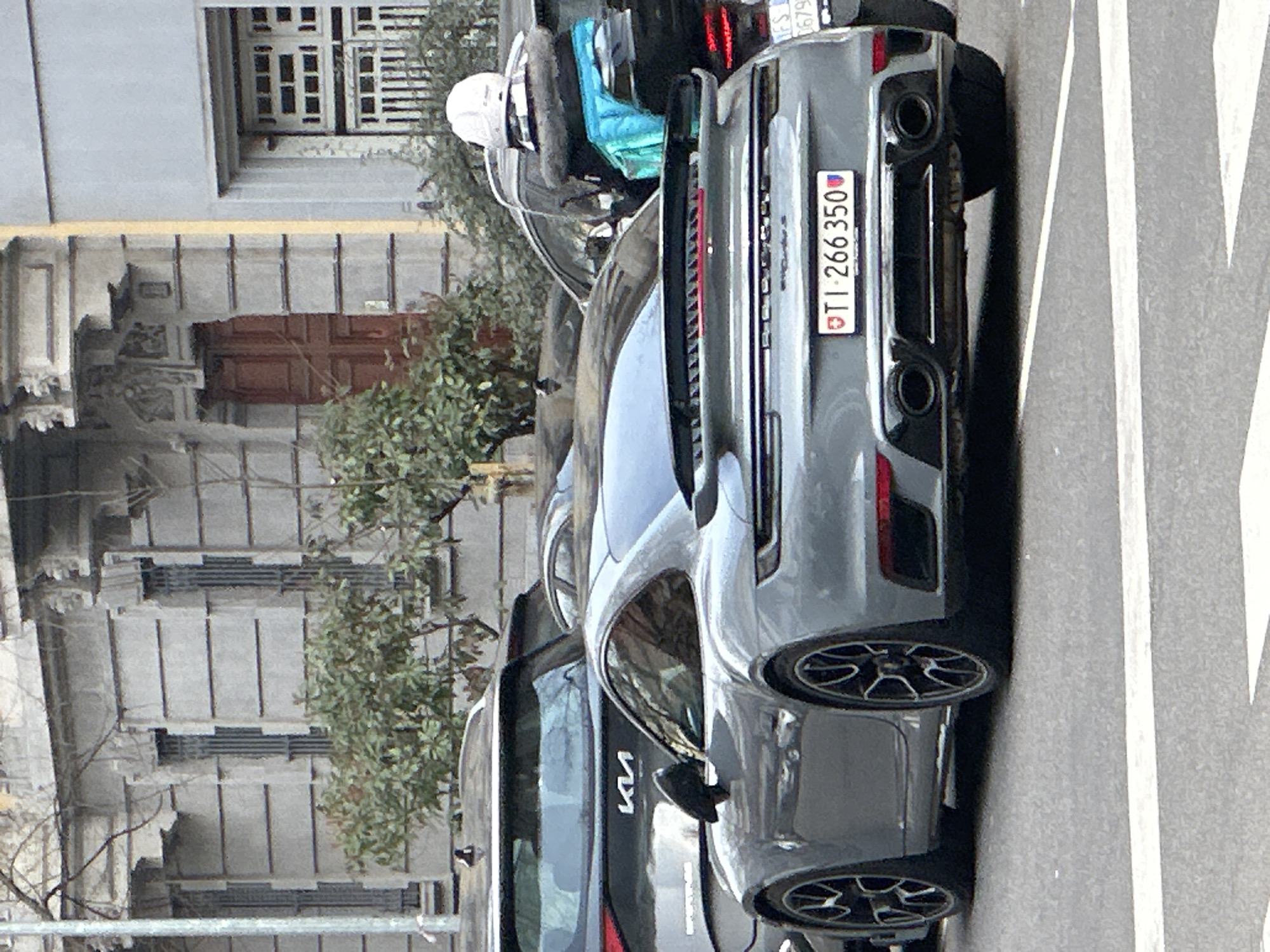 The image displays an urban street scene in Milan, Italy, under daylight conditions. A grey Porsche 911 (992 generation) sports car is the central subject, viewed from its rear-three-quarter angle, facing away from the camera. The vehicle has a prominent rear spoiler and dual oval exhaust pipes. Its license plate is Swiss, displaying "TI 266 350". Partially visible behind the Porsche is a dark grey Kia SUV. To the immediate right of the Porsche, a dark vehicle, possibly a scooter, is present, with a white helmet and a bright blue jacket secured on its rear. The background features two multi-story buildings typical of Milanese architecture. The left building has a light grey, horizontally banded facade with ornate stone detailing above a dark red wooden entrance door and several windows fitted with vertical bars. The adjacent building on the right is a simpler light grey, featuring a large window with a white grid pattern. Green foliage from bushes and trees is visible between the vehicles and in front of the buildings. The vehicles are positioned on an asphalt road, with white painted lines visible in the foreground.