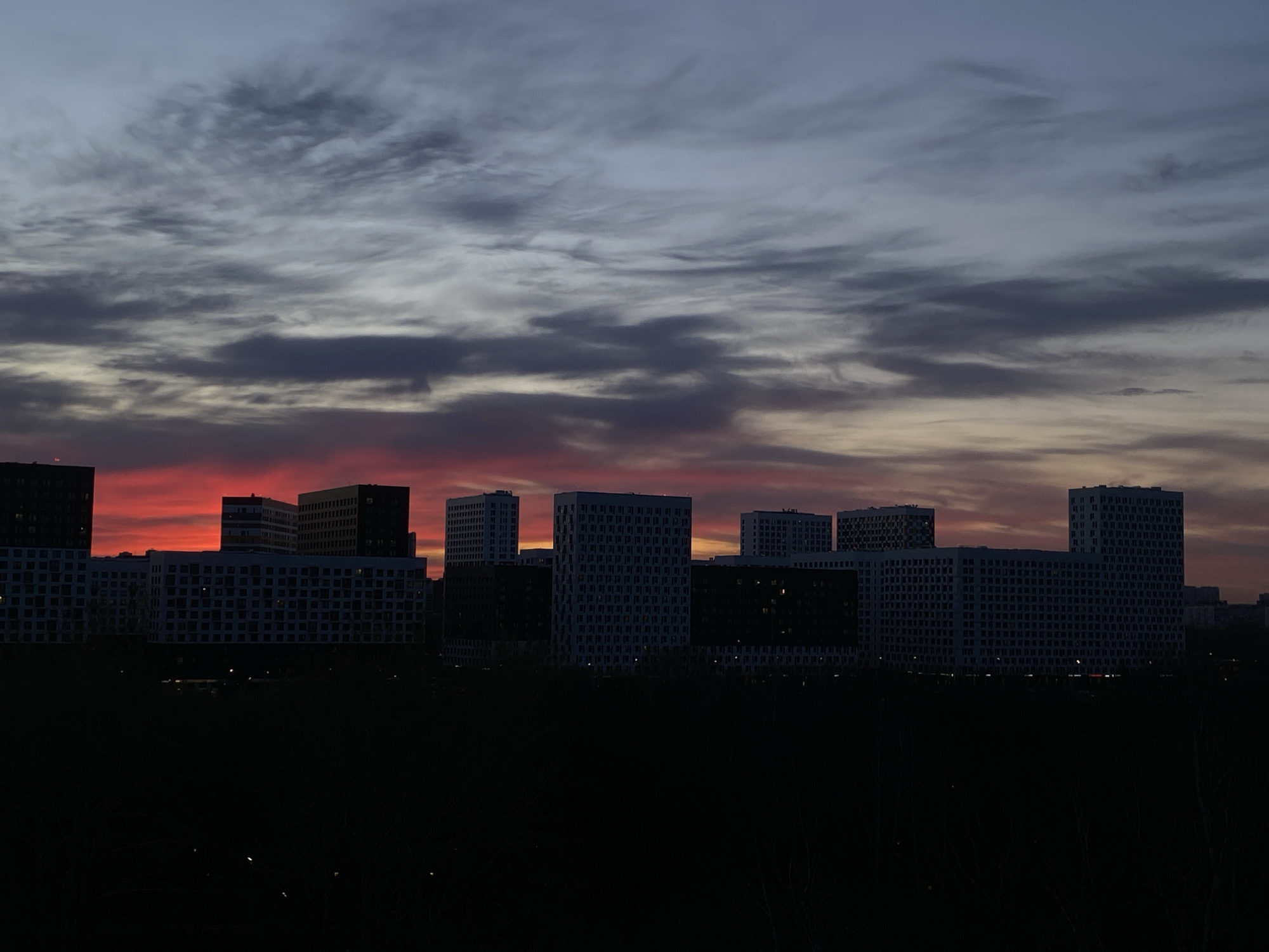 The image shows a cityscape at dusk or dawn. In the foreground is a dark, obscured area likely filled with trees or vegetation. Above, silhouetted buildings of varying heights stand against a dramatic sky. The sky features a gradient of colors, transitioning from dark grays and blues above to a band of bright red and orange near the horizon, suggesting either sunrise or sunset. The buildings appear to be modern residential or commercial structures with a grid-like pattern of windows. The overall impression is one of urban development contrasted against the natural beauty of a colorful sky. Location is in Moscow, Russia.