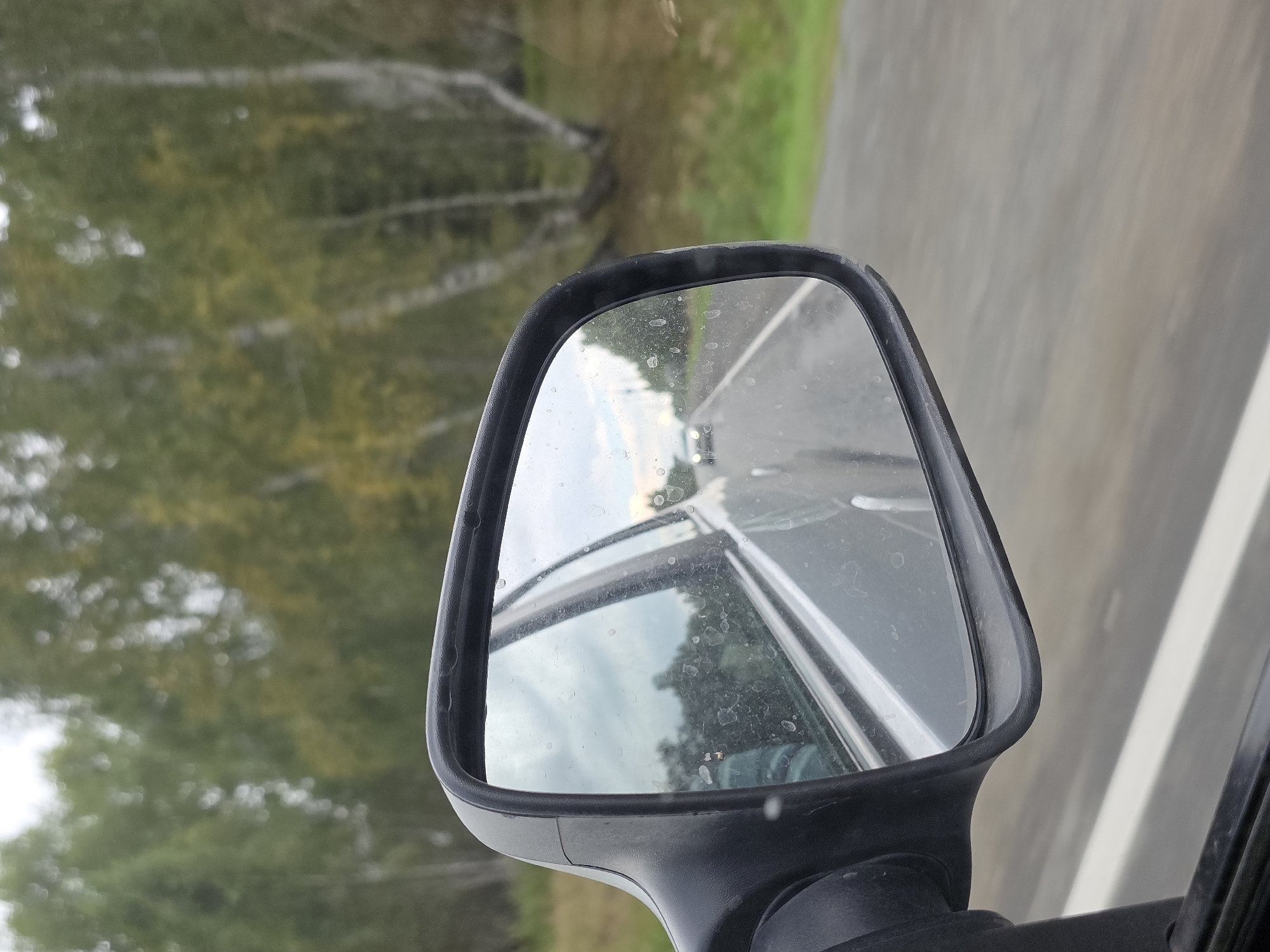 The image shows a close-up of a car's side mirror reflecting a road and trees. The mirror itself is dirty with several water spots. In the reflection, a road stretches into the distance, with a white line marking the lane. Another car is visible in the distance on the road. The background of the reflection shows a line of trees, some with autumnal foliage, and a cloudy sky. The surrounding environment appears to be a rural or semi-rural area, possibly along a highway. There are no people visible in the image. The image does not provide any information that would indicate the location is in Karsy, Russia.