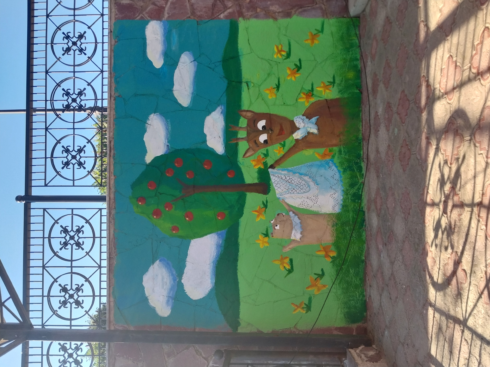 An outdoor mural is painted on a stone wall. The mural depicts a landscape with a green grassy field, a tree with red apples, a blue sky with white clouds, and yellow flowers. In the foreground, two cartoon animals, a brown bear and a deer, are interacting. The bear is wearing a white lace collar and the deer is wearing a blue bow. The deer is holding up a piece of white lace. The mural is located in the city of Orenburg, Russia, and is likely a part of a playground or public space. The ground in front of the mural is paved with decorative tiles, and shadows from a nearby fence are cast across the ground.