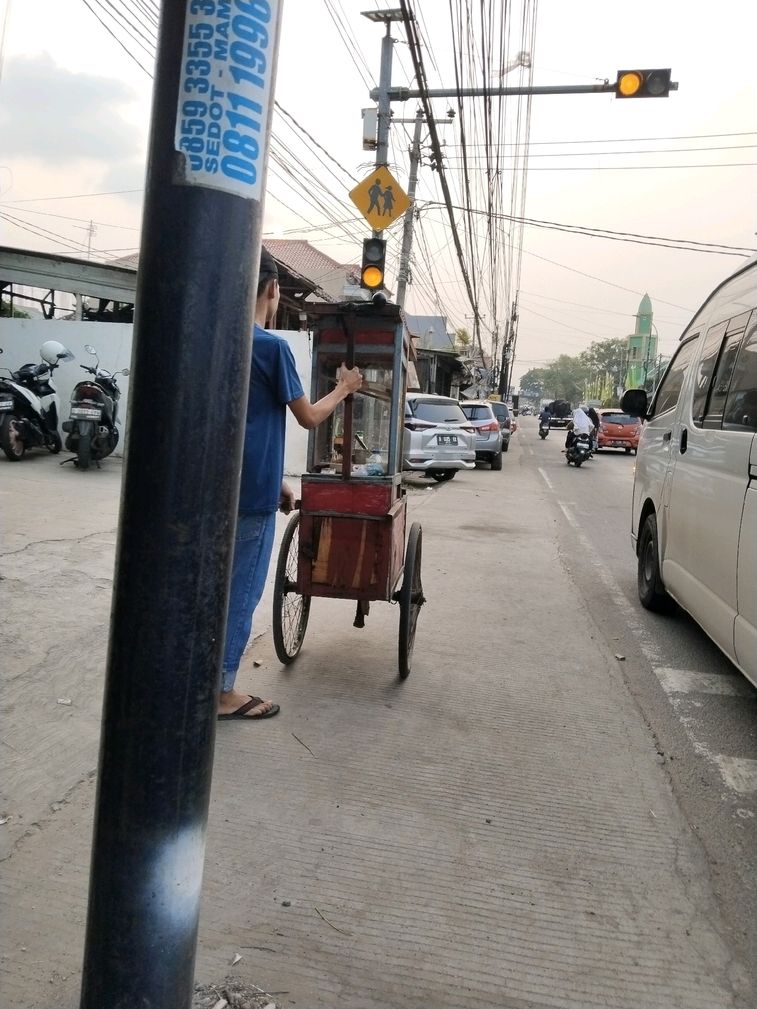 The image depicts a street scene in Serang, Indonesia, likely during daytime with overcast skies. In the foreground, a dark pole with white markings is partially visible on the left. To the right of the pole, a person wearing a blue t-shirt and jeans pushes a red cart with two wheels, possibly a food cart. The cart has a wooden frame and metal accents. Behind the cart, several motorcycles are parked. Across the street, there is a white van and other vehicles, including cars and motorcycles, on the road. Traffic lights and utility poles with a dense network of wires are prominent in the background. A yellow diamond-shaped pedestrian crossing sign is visible above a traffic light. Further down the street, a green dome, likely belonging to a mosque, can be seen amidst trees and buildings. The pavement is concrete with visible lines and some debris.