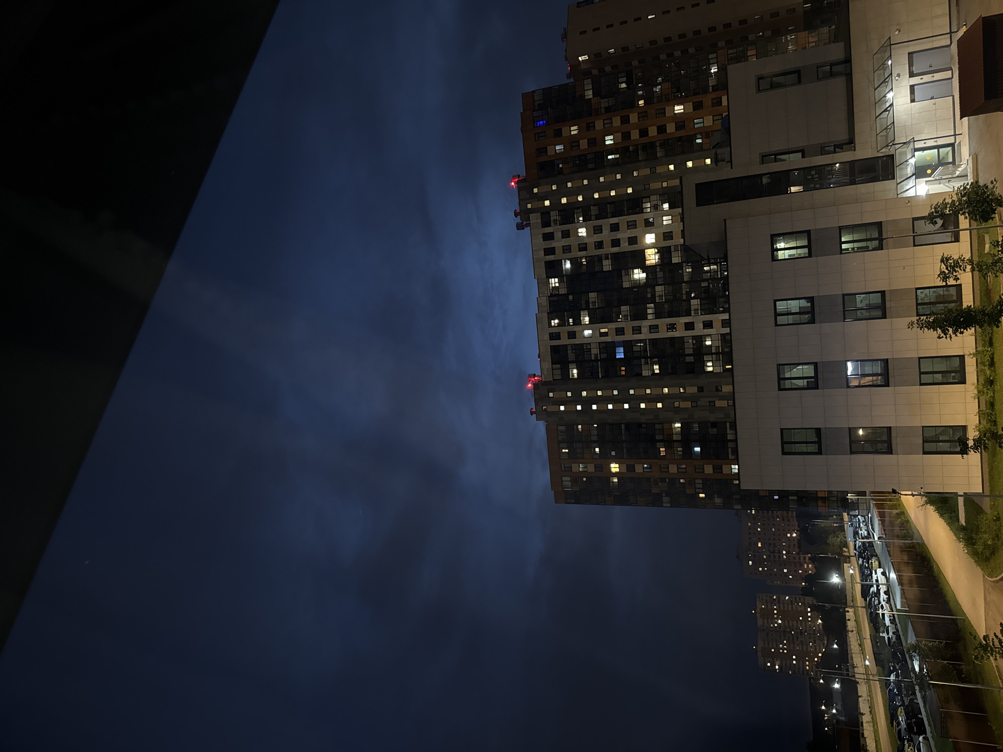 This is a night-time photograph taken in Vatutinki, Russia, capturing a modern urban scene dominated by tall residential buildings. The image is oriented vertically, with the buildings extending upwards on the right side of the frame and the night sky filling the majority of the left.
The primary subjects are two large, multi-story apartment complexes. The building in the foreground is a striking architectural piece with a mix of materials, including light-colored panels and darker sections with numerous lit windows, suggesting occupancy. The building behind it is taller and has a more uniform facade, also illuminated by many windows, creating a grid-like pattern against the dark sky. Red lights are visible at the top of one of the buildings, likely indicating aviation warning lights.
The sky is dark blue and appears cloudy, with some faint streaks suggesting atmospheric haze or light pollution. The overall atmosphere is that of a quiet, residential urban environment at night. Streetlights are visible in the distance, illuminating a parking lot filled with cars and a pathway. The presence of trees and a grassy area suggests an attempt at integrating green spaces within the urban development.
The time of day is clearly night, and the weather appears calm, with no indication of rain or strong winds. The illuminated windows suggest that this is a residential area where people are going about their evening routines. No people are directly visible, and the focus is on the architecture and the nocturnal urban landscape. There is no visible text in the image.