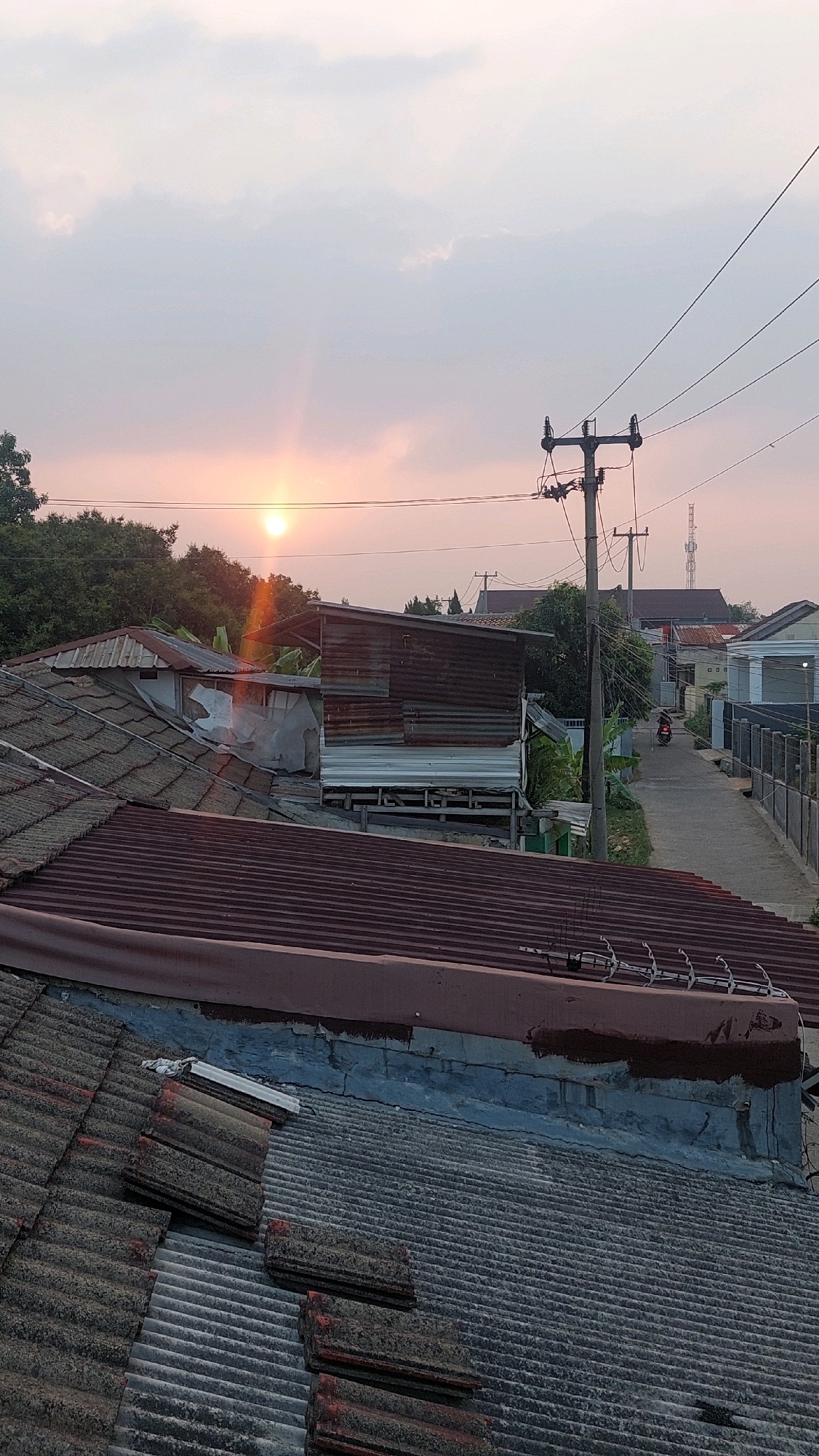 The image captures a twilight scene in Bojong Gede, Indonesia, with the setting sun casting a soft, orange glow across the sky. The foreground is dominated by the tiled roofs of several houses, some appearing weathered and discolored. Beyond these roofs, a narrow street winds through the neighborhood, bordered by houses and trees. A single motorcyclist is visible on the street, heading away from the viewer. Electrical poles with wires are prominent features of the urban landscape, crisscrossing the sky. The overall impression is one of a modest residential area at the close of day.