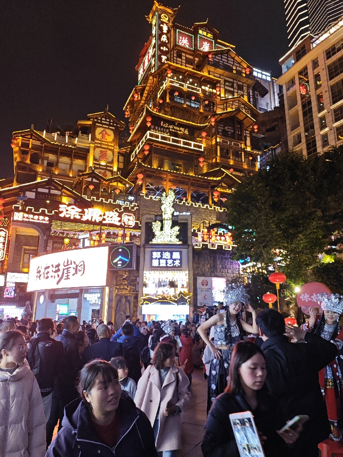 The image presents a densely crowded night scene in the Hongyadong complex, located in Yuzhong District, Chongqing, China. The prominent multi-tiered structure, characterized by traditional Chinese architecture with upturned eaves and wooden frameworks, is extensively illuminated with warm golden and white LED lights, outlining its details. Multiple signs with Chinese characters are visible on the building, including "洪崖洞" (Hongyadong), "金寨" (Jinzhai), "重庆" (Chongqing), and "好吃街" (Delicious Food Street). Red traditional Chinese lanterns hang from various sections of the architecture.
Below the main structure, a large rectangular electronic sign displays "我在洪崖洞" ("I am in Hongyadong") in white Chinese characters. Adjacent to it, another large sign advertises "郭浩晶 雕塑艺术" (Guo Haojing Sculpture Art), positioned below a brightly illuminated, intricate sculpture resembling a mythical figure situated on a crenellated stone wall. To the upper right, a modern skyscraper with grid-patterned windows is partially visible, contrasting with the traditional complex. A large tree with dark foliage is on the right side of the frame.
The foreground and midground are filled with a large crowd of pedestrians, primarily East Asian individuals, navigating the bustling area. Most are dressed in casual outerwear, such as jackets and coats. On the right side, several individuals are adorned in elaborate festive costumes, including shimmering silver headpieces and patterned garments, appearing to be performers or street entertainers. One female in such attire smiles while adjusting her headpiece. Many people are engaged with their surroundings; several individuals in the foreground are visibly holding and interacting with smartphones. Red spherical objects or balloons, some with Chinese characters, are also visible among the crowd.