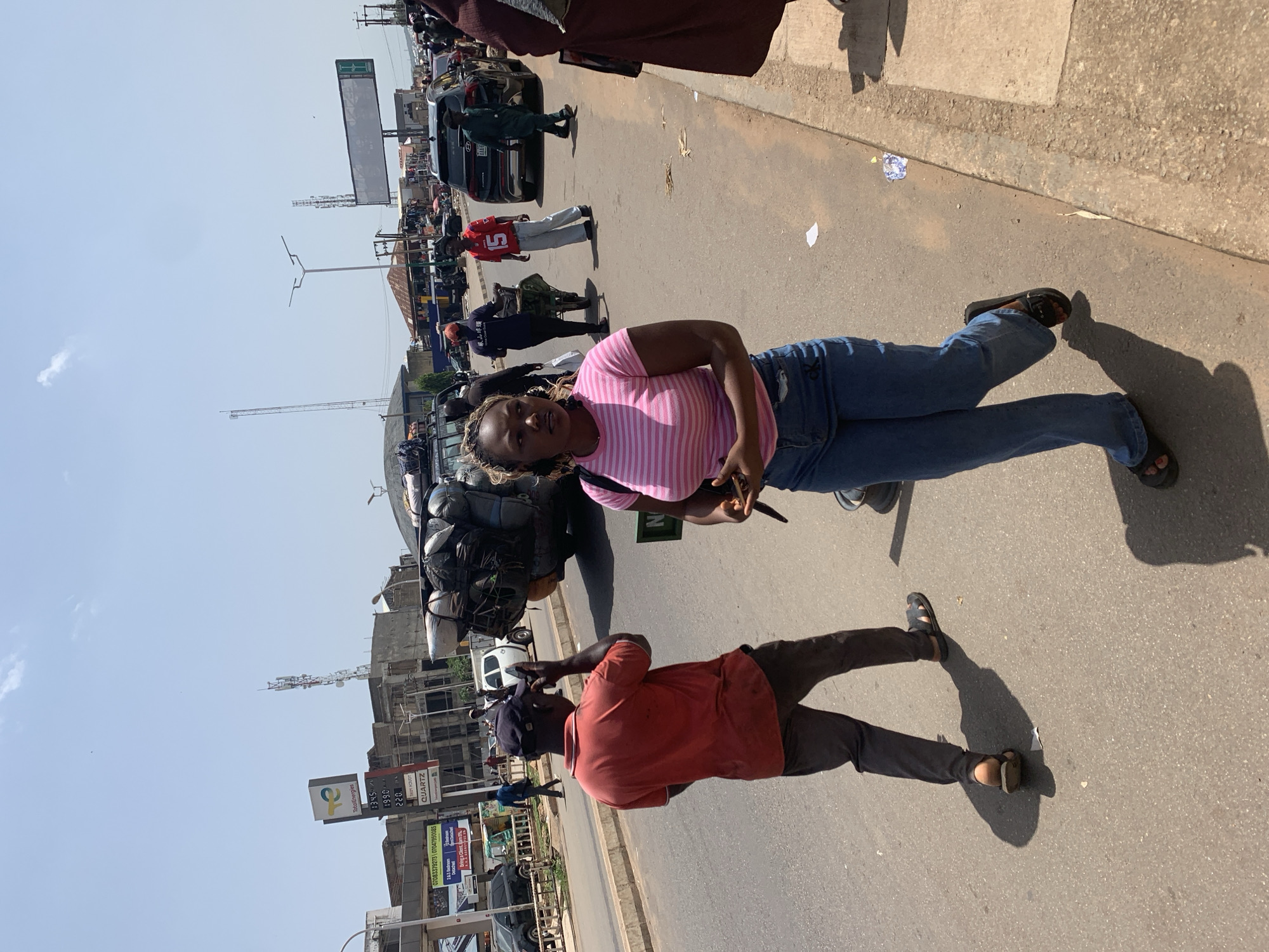 In Jos, Nigeria, on a sunny day, a woman in a pink and white striped t-shirt and blue jeans walks along the side of a busy road. She is holding a black object in her right hand. Behind her, a man in a red shirt and dark pants, wearing a dark cap, is walking while holding a phone to his ear. A vehicle overloaded with luggage is parked on the right side of the road. In the background, there are buildings, a gas station sign, traffic lights, and other vehicles, including a black SUV. People are seen walking on the sidewalk and in the street, and a man is pushing a cart. The environment suggests an urban setting with commercial activity.