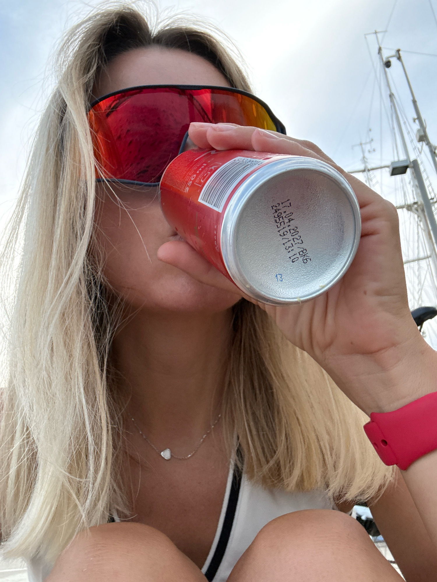 A person is drinking from a red can of Coca-Cola while on a boat. The person has blonde hair and is wearing red sunglasses, a silver necklace, and a bright pink watch. In the background, the masts of sailboats are visible against a cloudy sky, suggesting the boat is docked or sailing in a marina in Valencia, Spain.