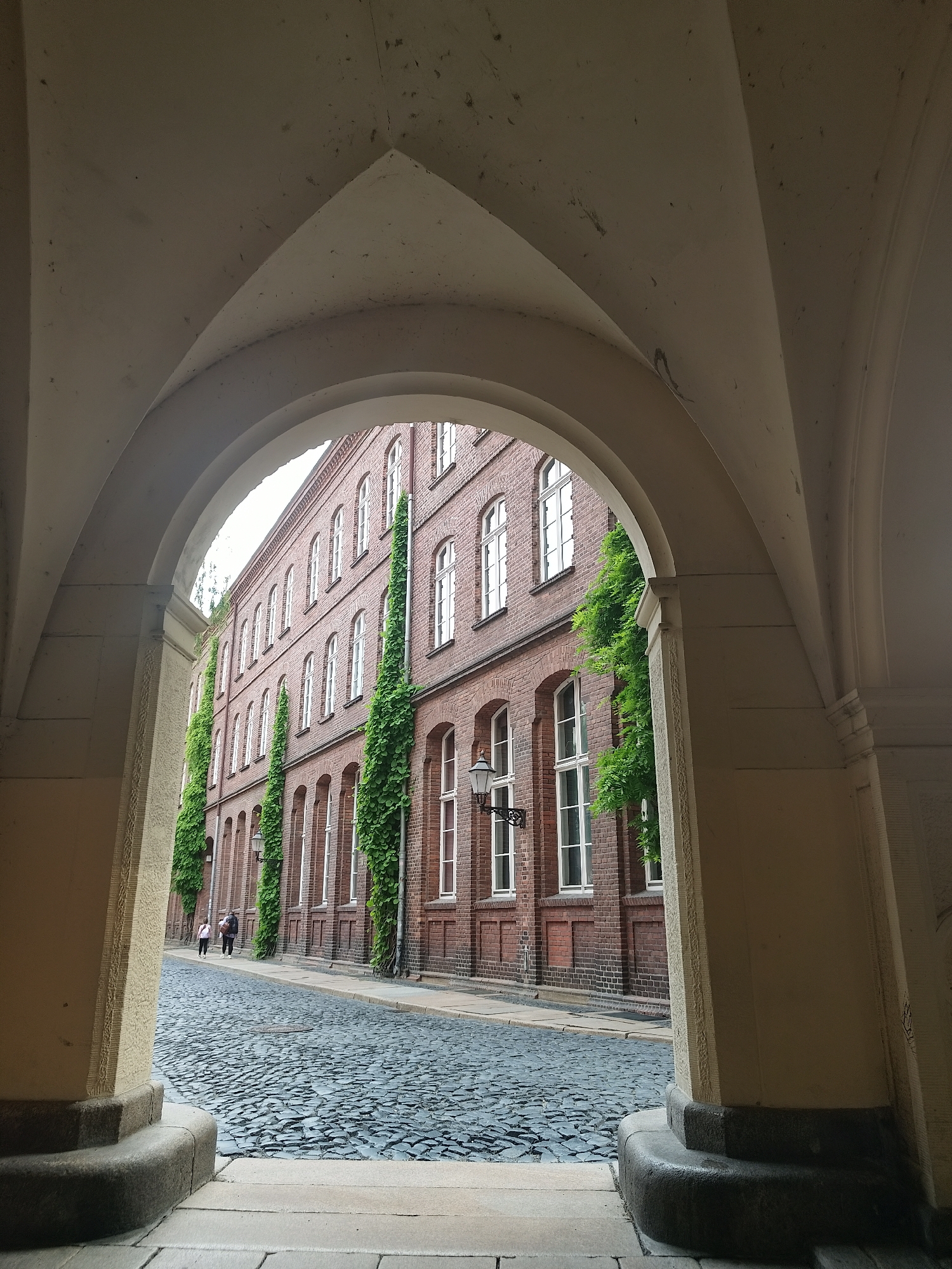 The image depicts a street scene viewed through an arched opening. The arch is constructed from light-colored stone, revealing a cobblestone street lined by a brick building. Green vines climb vertically along the face of the brick building. Two people are walking on the street in the distance. The photograph was taken in the city of Unknown, Unknown.