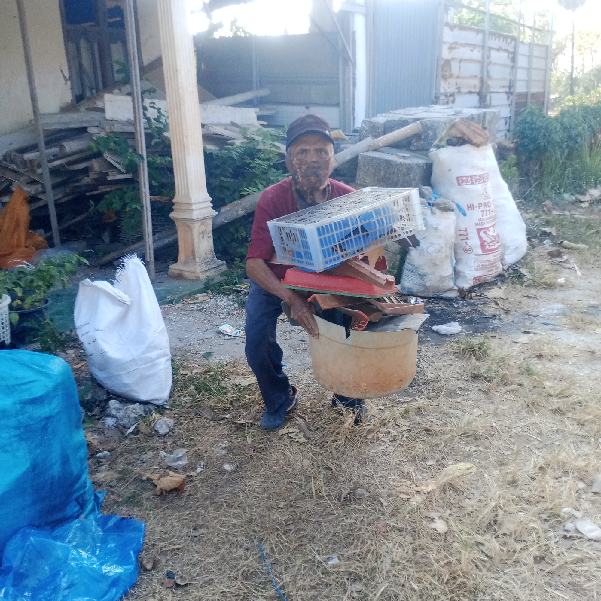 In Semanding, Indonesia, a man with a dark baseball cap and a maroon shirt stands outdoors, carrying a large tan bucket filled with debris. His face appears smudged with dirt or paint. In his hands, he holds a blue plastic crate overflowing with various items, including red plastic pieces and what appear to be roof tiles. The man is positioned on a dry, grassy area dotted with leaves and small debris. Behind him, several large white bags are visible, one of which has "HI-PRO 771" printed on it. The background shows a building with corrugated metal siding and piles of discarded materials, suggesting a construction or cleanup site. To the left, a large blue tarp is partially visible, lying on the ground. The overall environment is dry and somewhat unkempt, consistent with an outdoor area undergoing renovation or waste disposal.