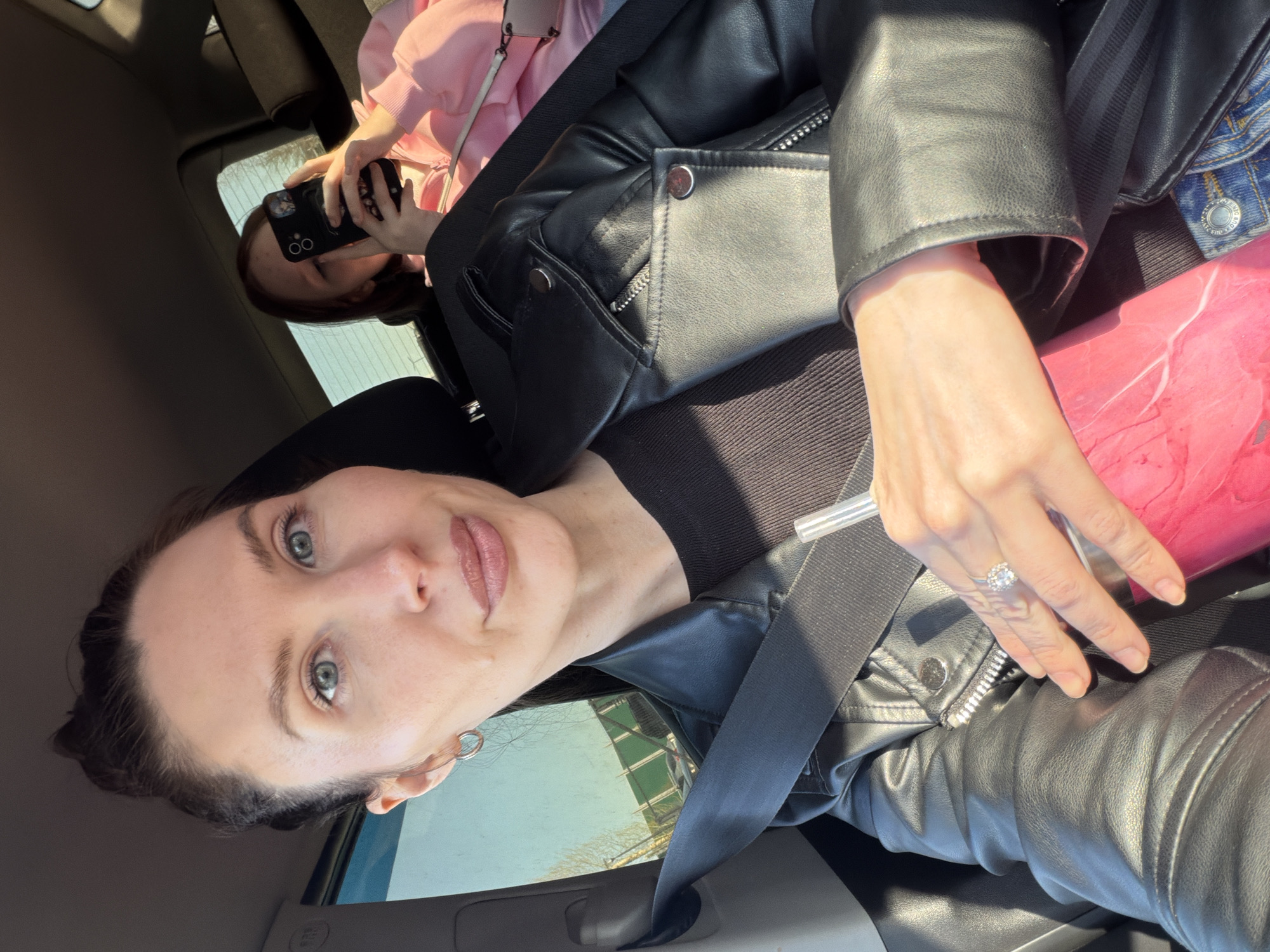 A medium shot taken from inside a vehicle shows a woman with dark hair and light eyes, wearing a black leather jacket and a black top, with a seatbelt across her body. She has a ring on her finger and is holding a pink cup. In the background, another person, a female, wearing a pink hoodie, is holding a black phone, presumably taking a photo. The scene suggests they are in a car, the window is visible with an outside view that shows trees and buildings with green accents. The exact location can not be determined, but it would be an urban environment in the city of Perm, Russia.