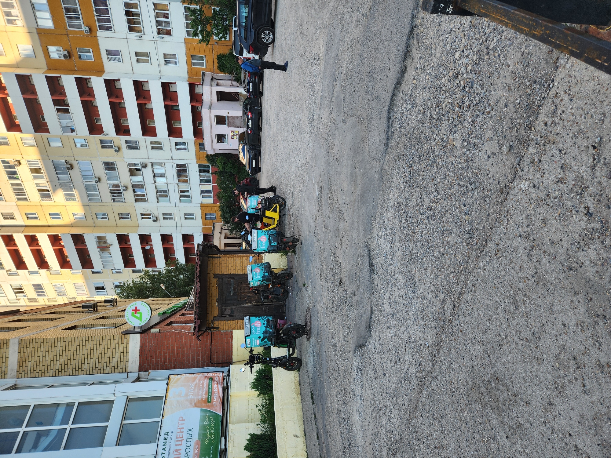 In the city of Vniissok, Russia, on a paved lot, a group of delivery couriers are gathered with their electric scooters. Five electric scooters are parked in a line, each equipped with large, insulated delivery boxes. Three of the boxes are labeled "OZON fresh" and feature a teal and white design. Two couriers, wearing black shirts, are standing near one of the scooters, possibly preparing for deliveries. A third courier in a blue jacket stands beside a black SUV in the background. Several parked cars are visible, including black sedans and SUVs, suggesting this area may be a pickup or staging point for delivery services. The background consists of a multi-story residential building with a light yellow and white facade, accented with red balconies. Adjacent to the residential building is a smaller brick structure with a dark wooden door. A sign with a red cross and green text, possibly indicating a medical facility, is mounted on the brick building. The ground is covered in gravel and cracked asphalt, with some patches of green vegetation visible along the edge of the lot.